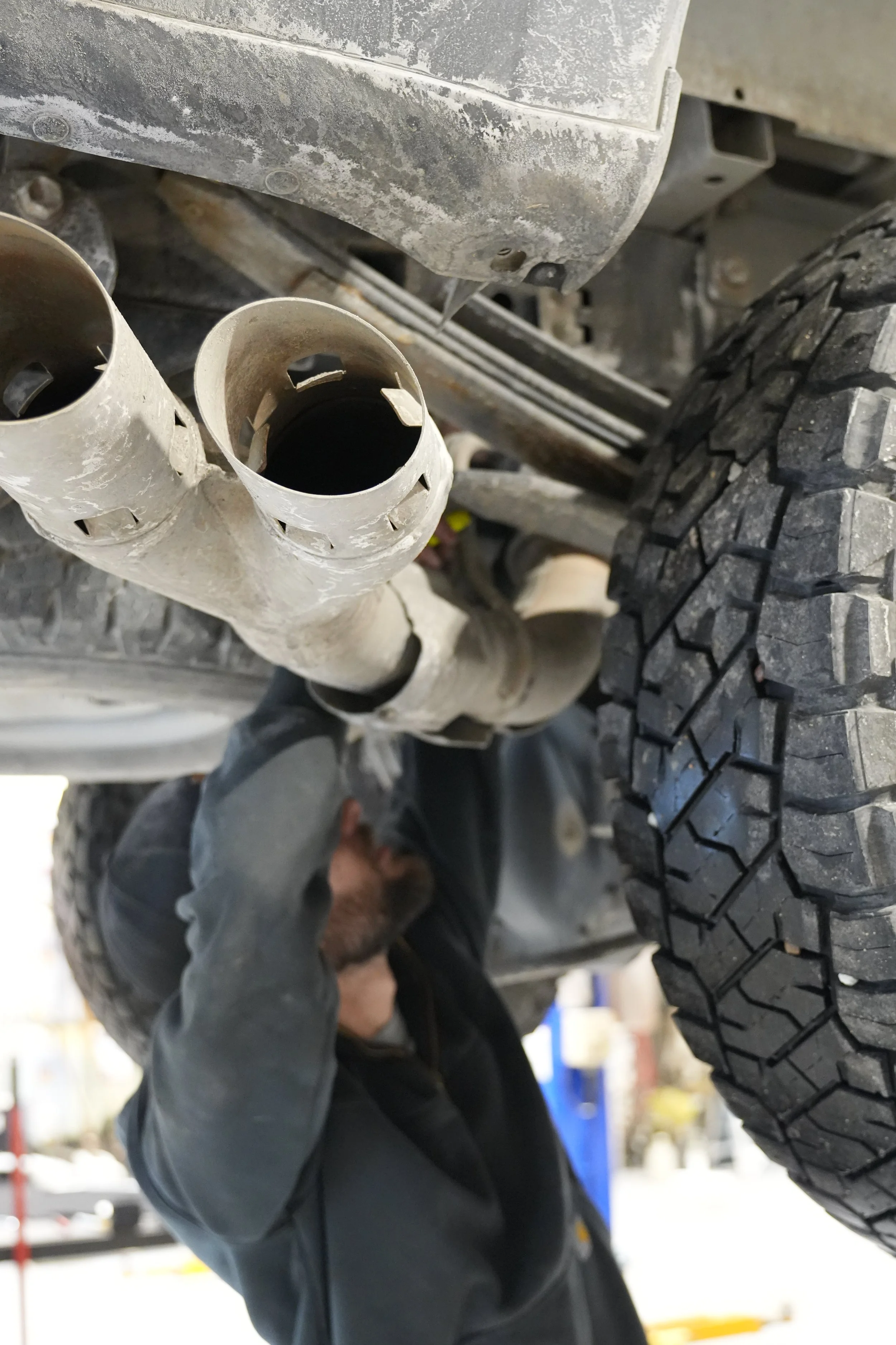 A mechanic inspecting or working on the underside of a vehicle, near the exhaust pipes and tire, wearing a black jacket and gloves in an auto repair shop.