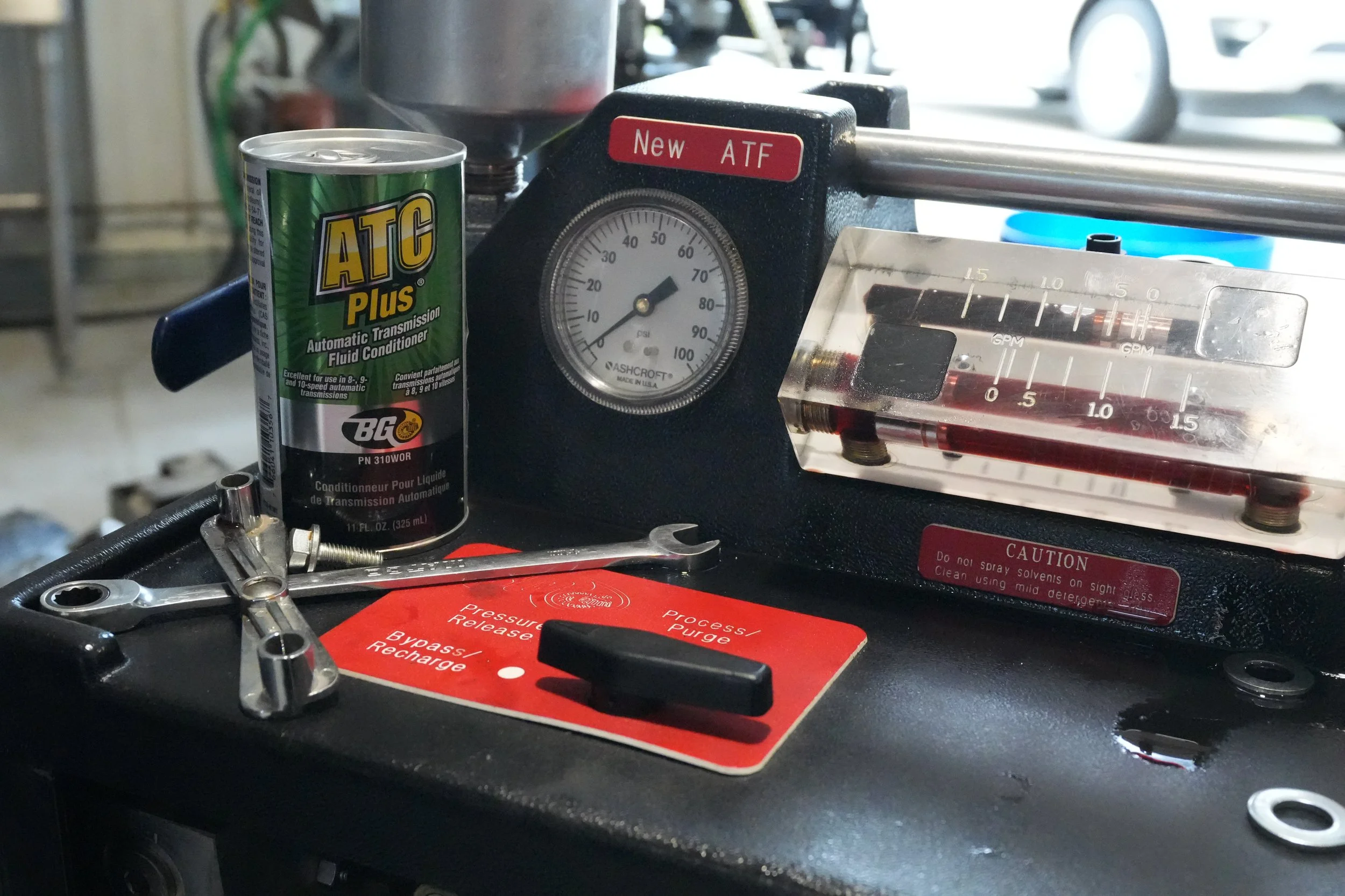 A can of ATC Plus automatic transmission fluid, a black mechanical device labeled 'New ATF,' a clear plastic gauge reading fluid levels, and a red warning label with instructions are visible on a mechanic's workbench. The background shows a garage with cars and tools.