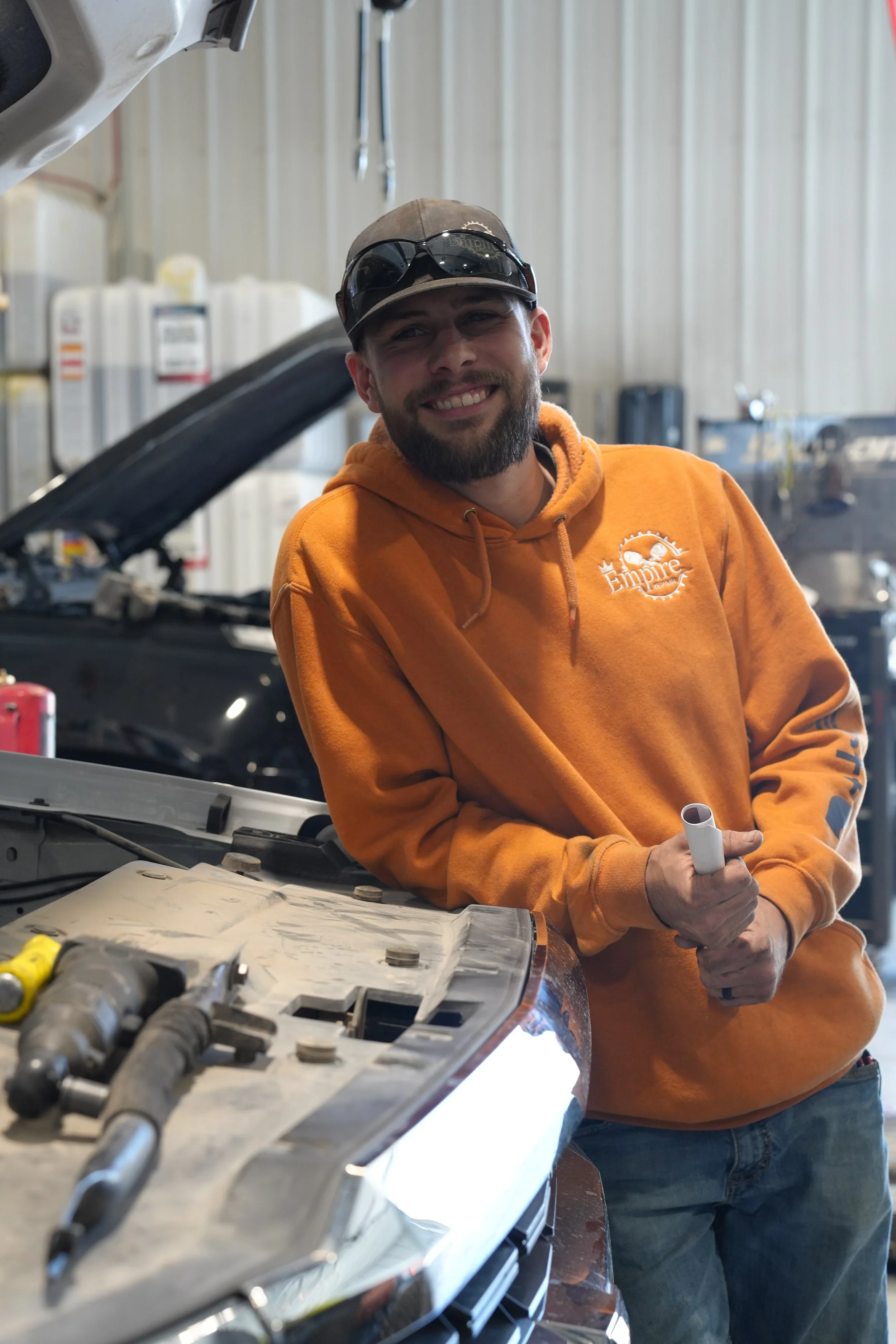 A man with a beard smiling and wearing an orange hoodie with 'Empire' logo, holding a tool, standing next to an open car hood in a workshop.