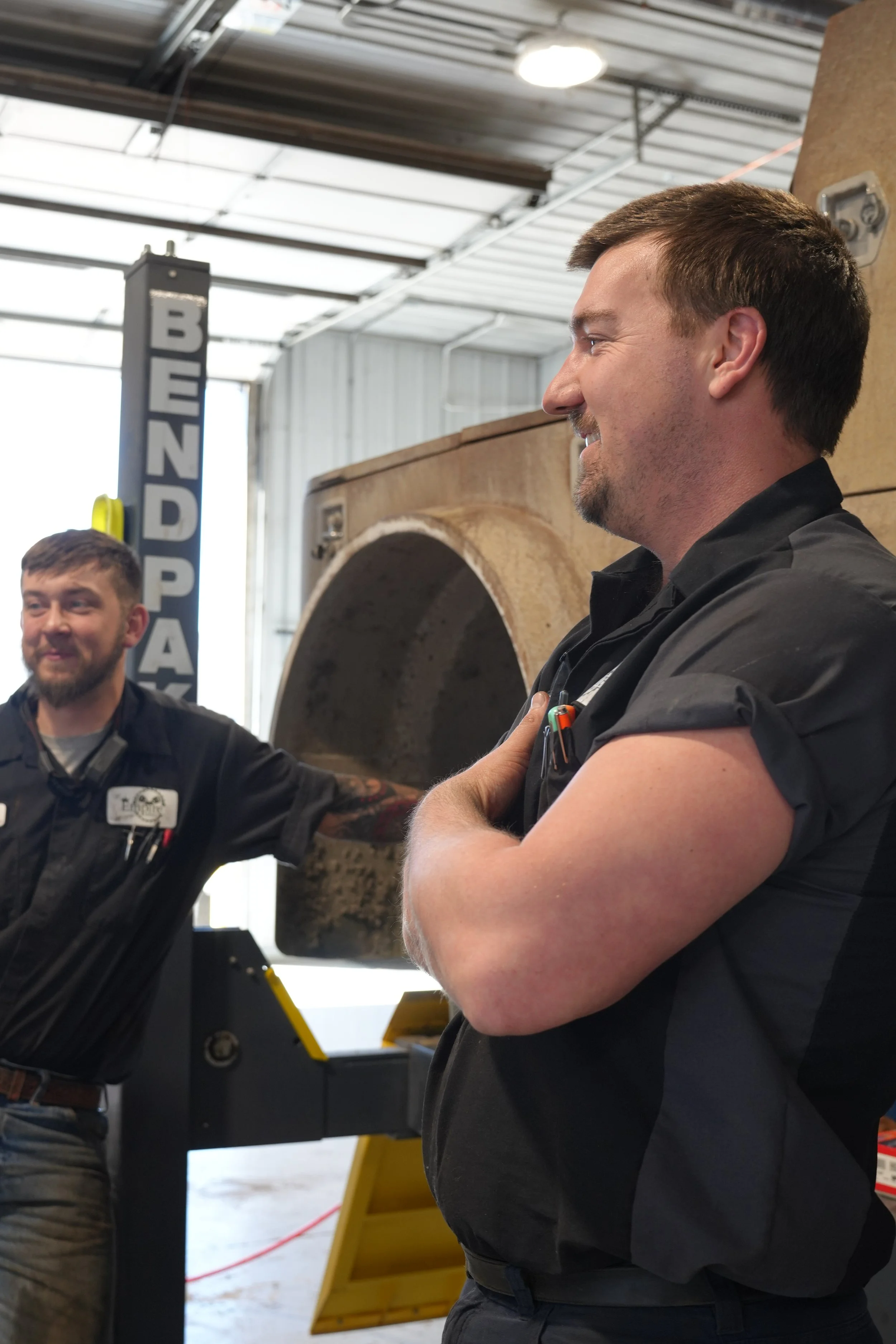 Two men in work uniforms inside a workshop or industrial setting. One is smiling with arms crossed, and the other is smiling and gesturing with his hand. There are industrial tools and machinery in the background, with a visible vertical sign that reads 'BEND PAR'S'.