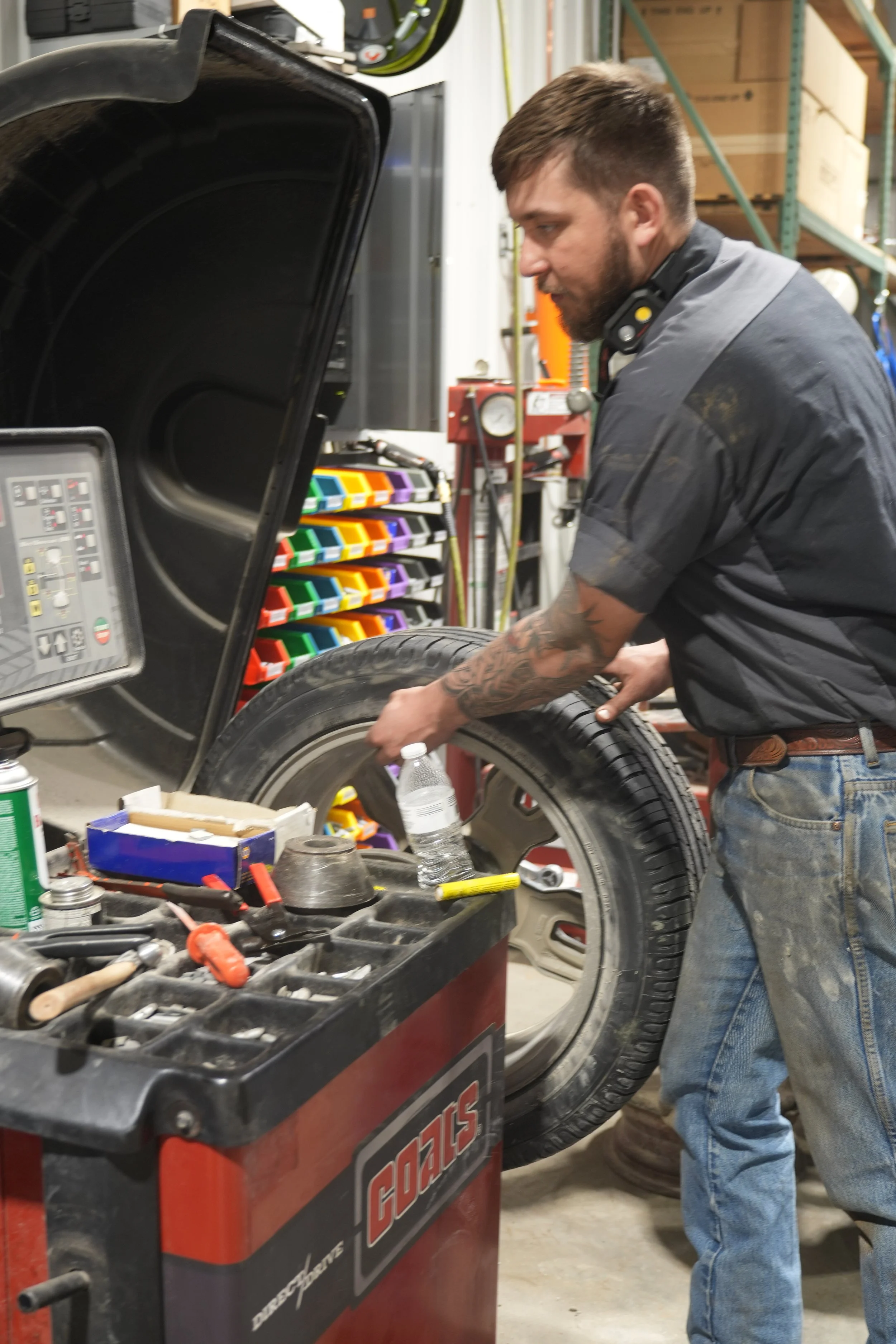 A man working on a tire in an industrial workshop or garage, surrounded by tools and equipment, with shelves containing boxes and a color-coded organizer in the background.