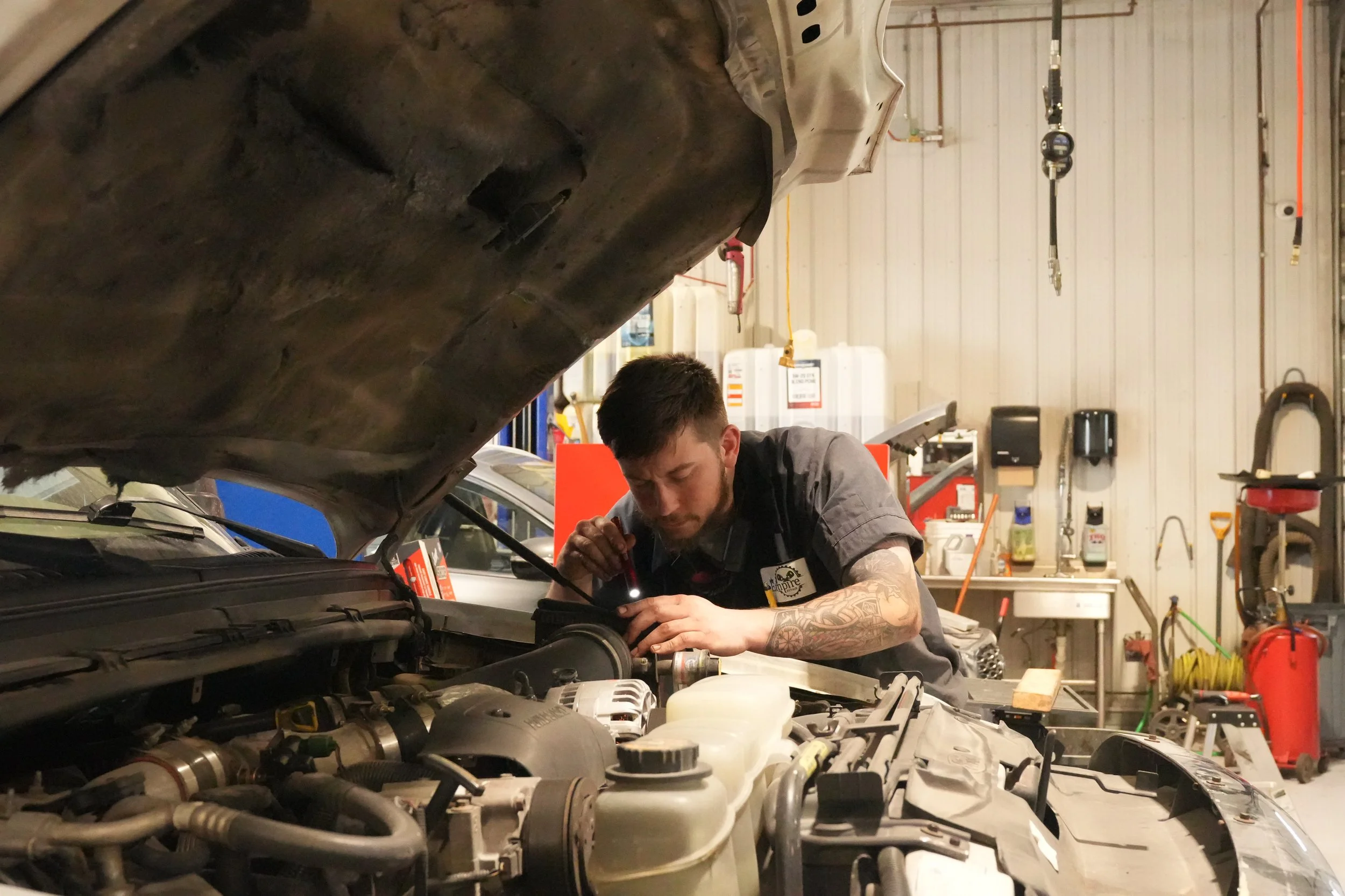 A mechanic working on a car engine in a garage, leaning over the open hood of the vehicle, with various tools and equipment in the background.