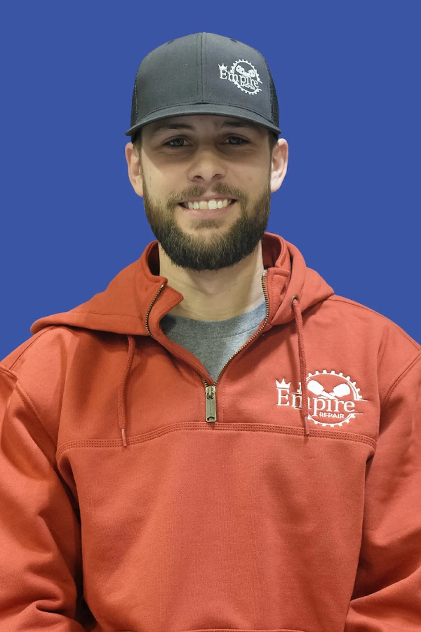 A man smiling, wearing a black cap and a red hoodie with a logo that says 'Empire Repair' on the chest, against a solid blue background.