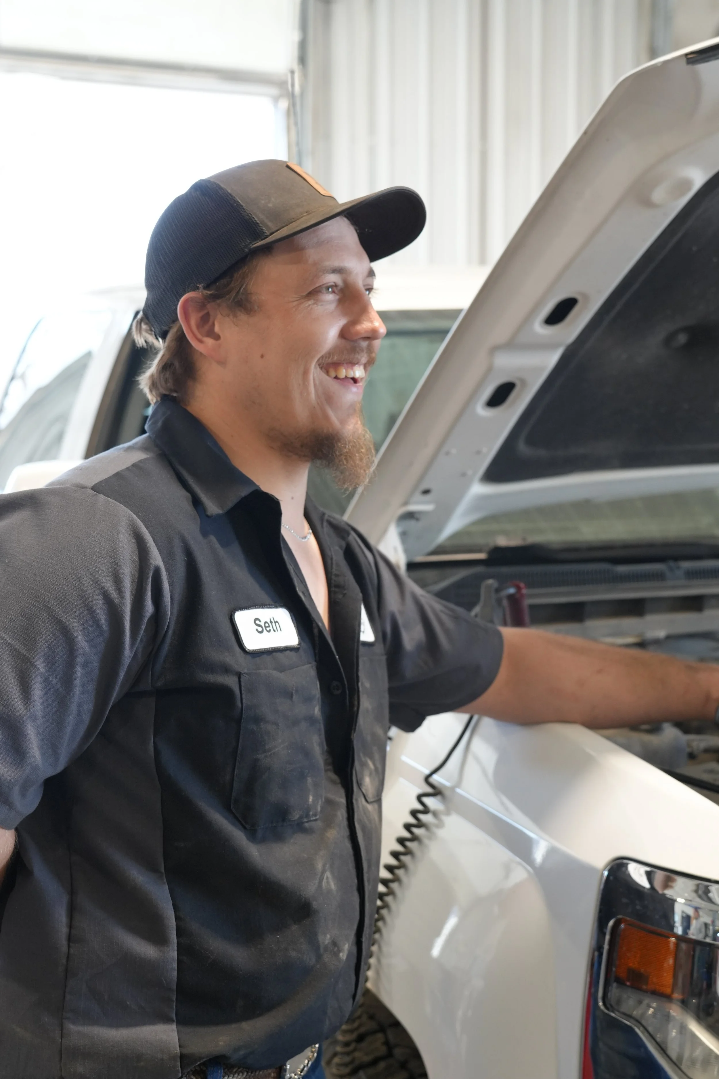 A man wearing a black shirt and a black and gray baseball cap, smiling as he works on a white vehicle with its hood open in a garage.
