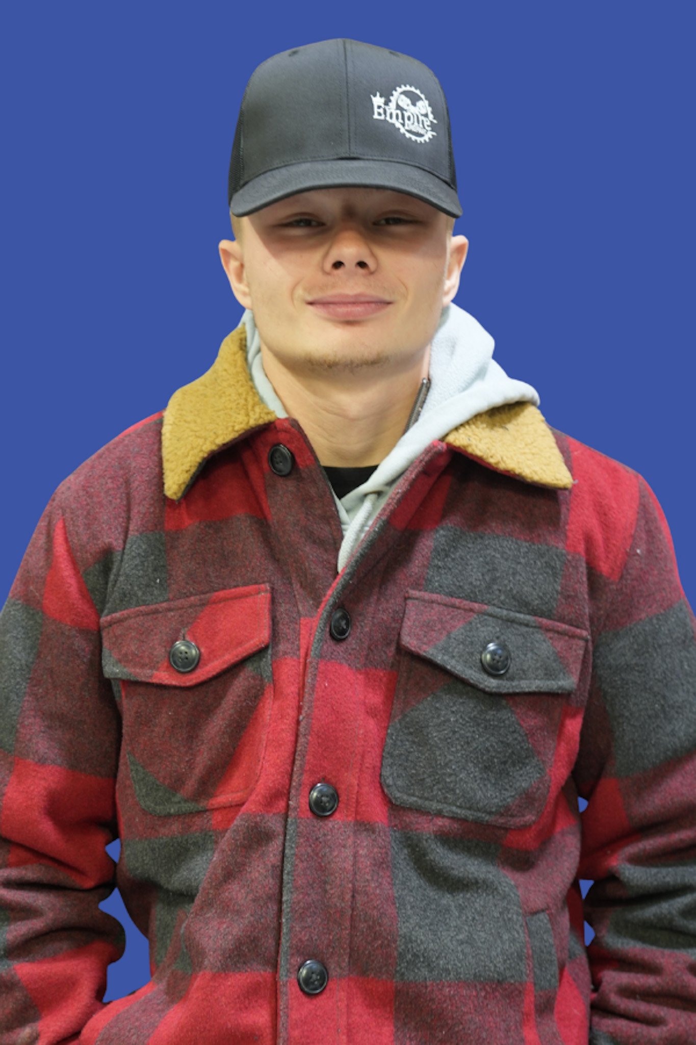 A young man wearing a dark baseball cap with a logo, a hoodie, and a red plaid jacket standing against a solid blue background.