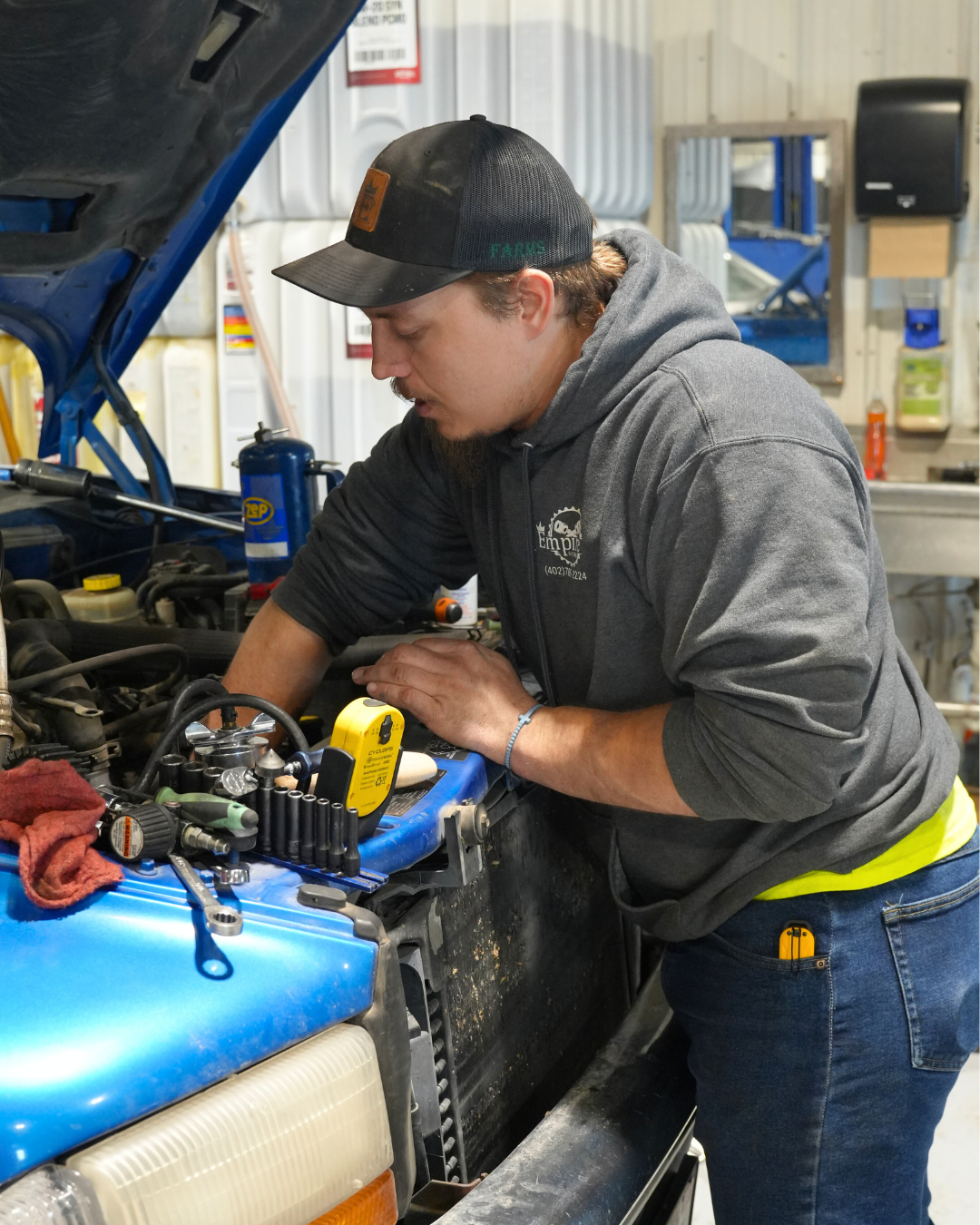 A man working on a blue vehicle engine in a garage or workshop, wearing a gray hoodie and a black cap.