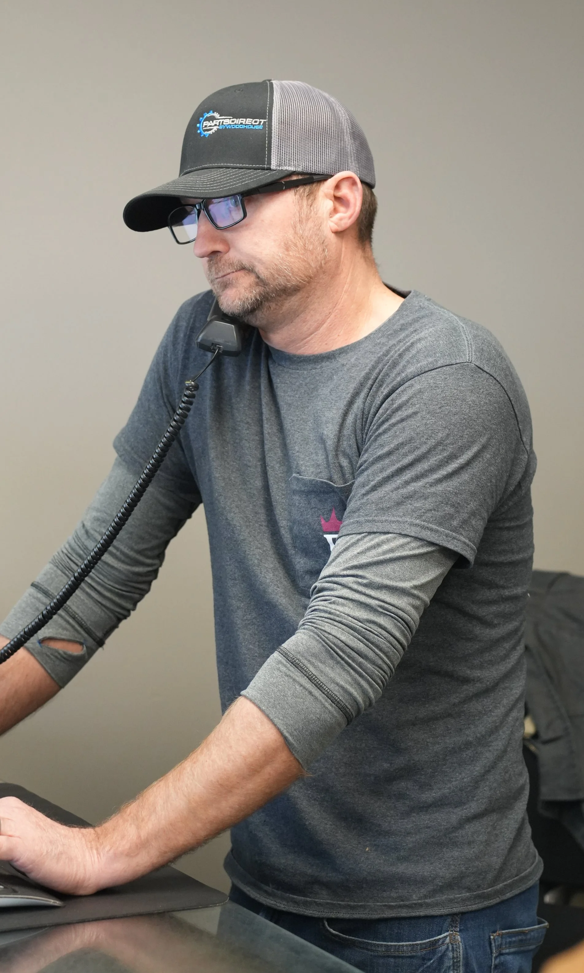 A man wearing glasses and a gray and black baseball cap, with a logo on the front, is looking at a computer screen. He is dressed in a gray long-sleeve shirt and is standing at a desk with a computer mouse and keyboard.