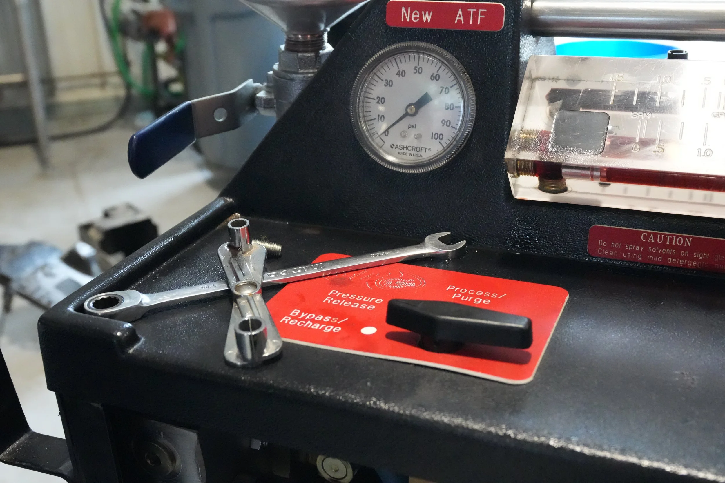 A pressure test gauge system with a pressure gauge reading approximately 65 psi, a red and white label reading 'New ATF', wrenches, and a red caution label with instructions for handling fluids.