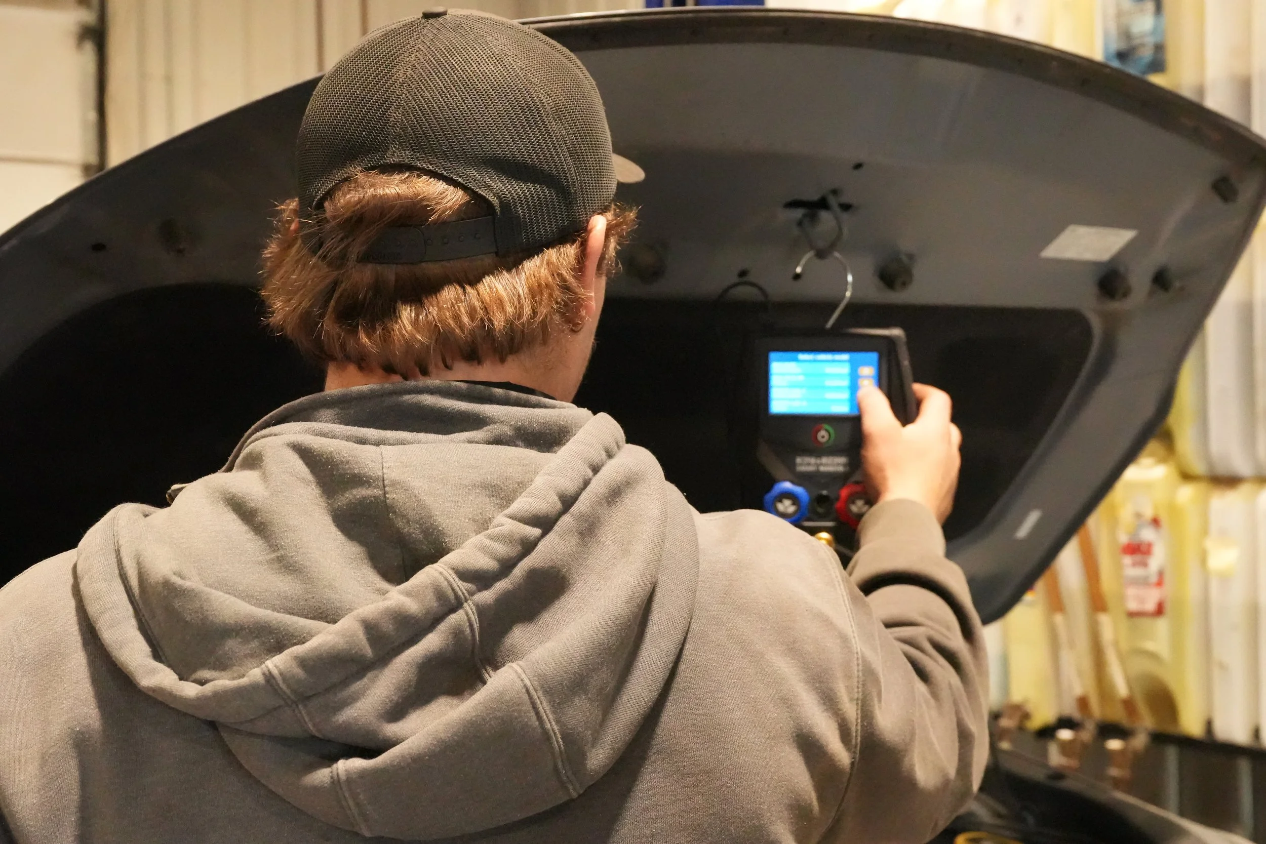 A person with brown hair wearing a gray hoodie and black cap is working with a diagnostic scanner in a garage or auto repair shop.