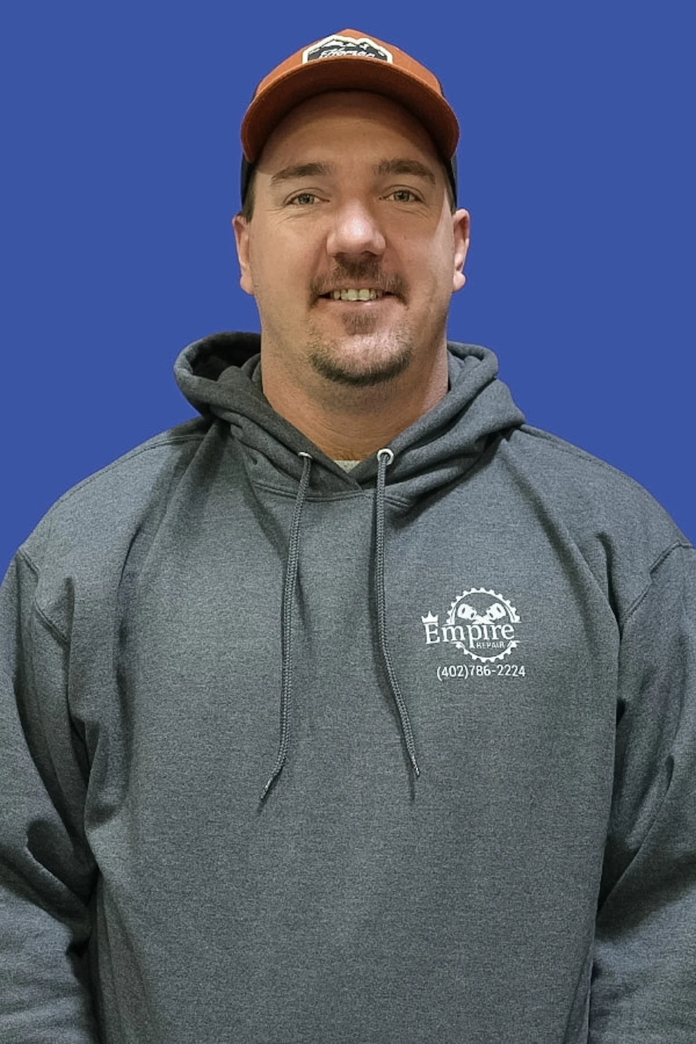 A man smiling in a gray hoodie and a baseball cap against a blue background.