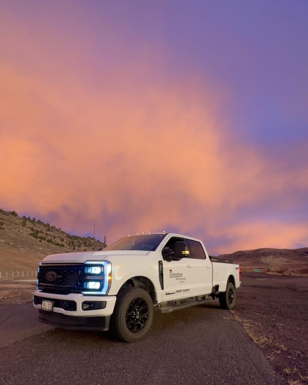 A white Ford pickup truck parked on a dirt road with a sunset sky showing pink and purple clouds in the background.