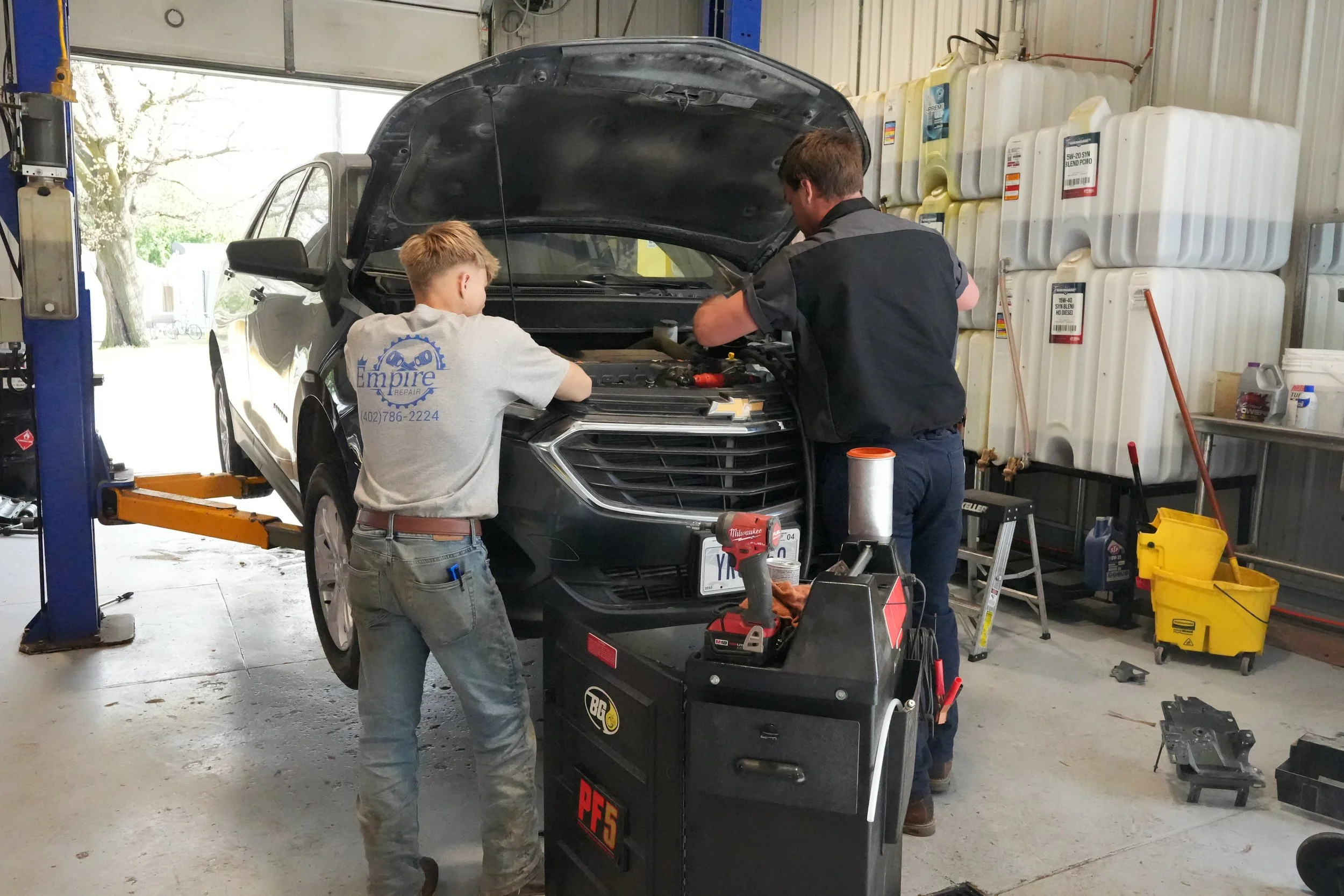Two men who are mechanics, one in a gray short sleeve shirt with a blue empire repair logo and the other in a gray short sleeve mechanic shirt stand in front of a black chevy SUV with the hood up looking into the engine.