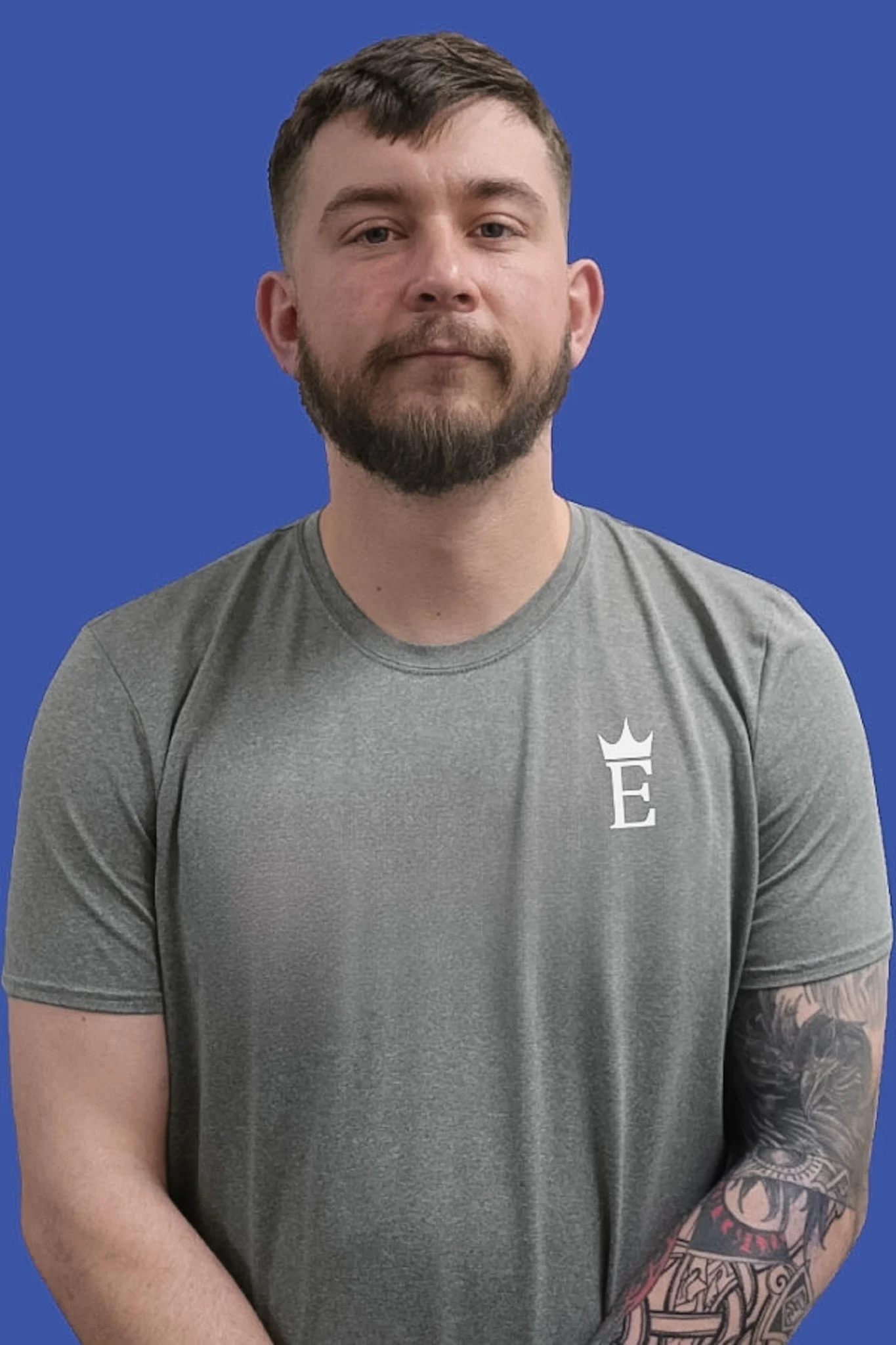 A young man with short brown hair and a beard, wearing a gray t-shirt with a white emblem on the chest, standing against a solid blue background. Part of a tattooed arm is visible.