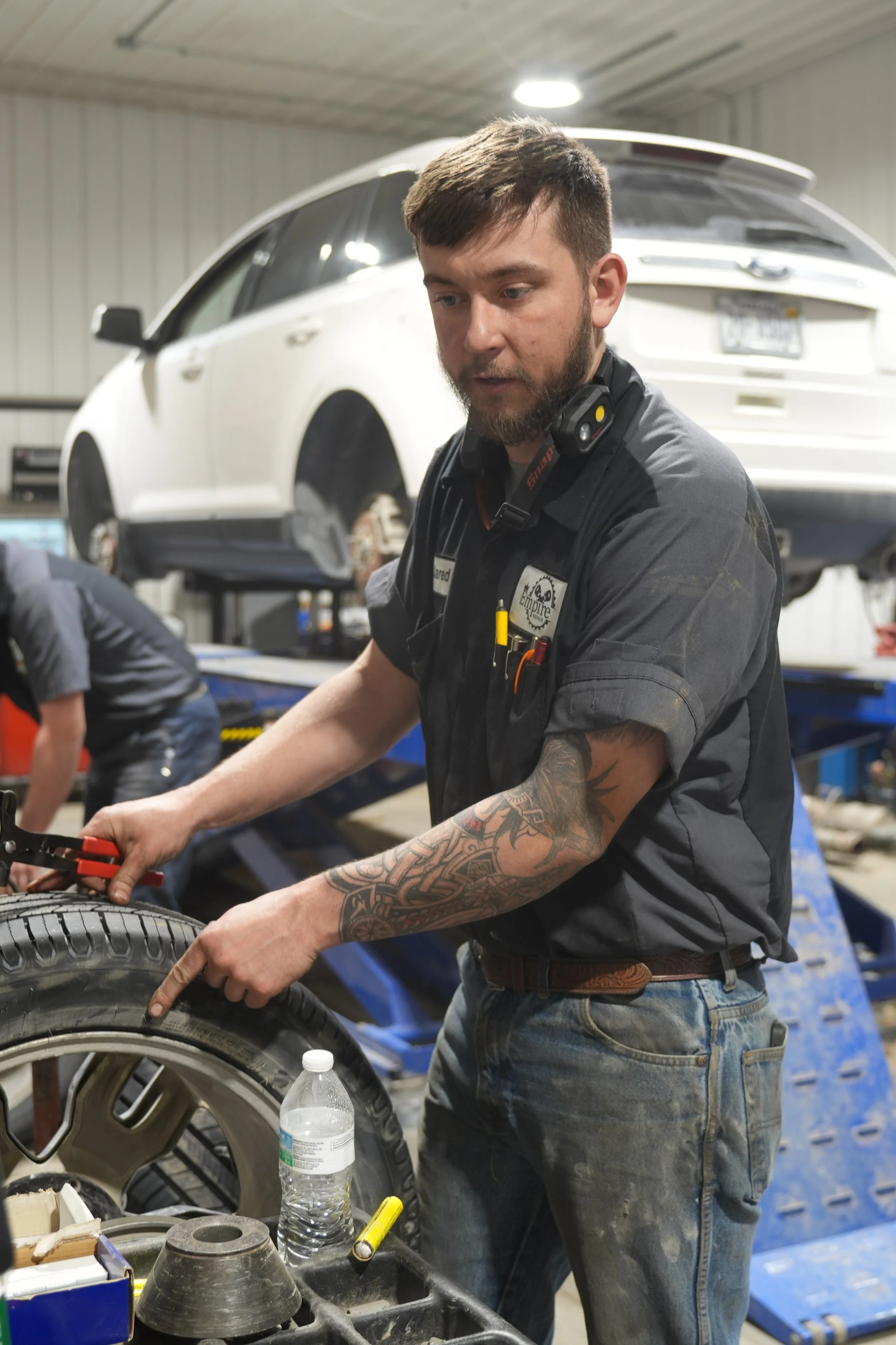 A man working on a car tire in a garage, with a white SUV on a lift in the background.