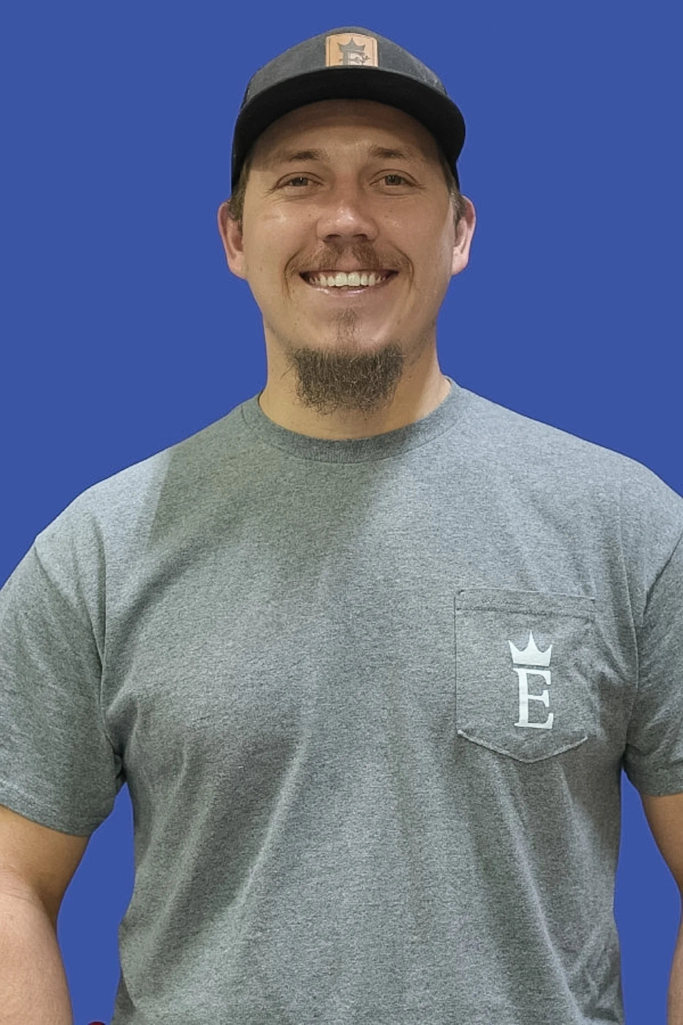 A smiling man wearing a gray t-shirt with a pocket that has a white logo and a dark baseball cap with a patch on the front, standing against a blue background.