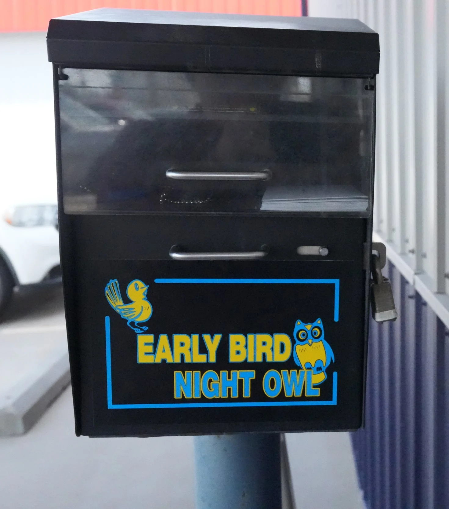 A black kiosk with a transparent top section, featuring a yellow and blue sticker with a bird and an owl, and the words "Early Bird Night Owl" in yellow and blue.
