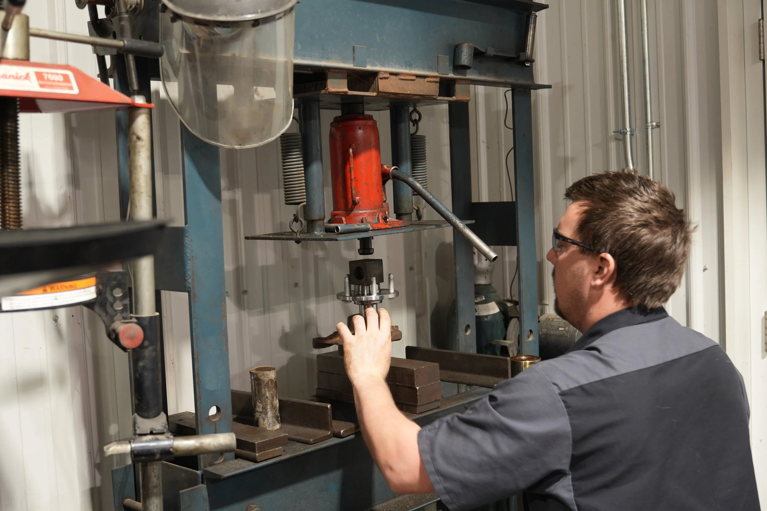 A person working in a machine shop, adjusting a piece of equipment on a metal machine with gears, springs, and a hydraulic red cylinder, in a workshop with a corrugated metal wall.