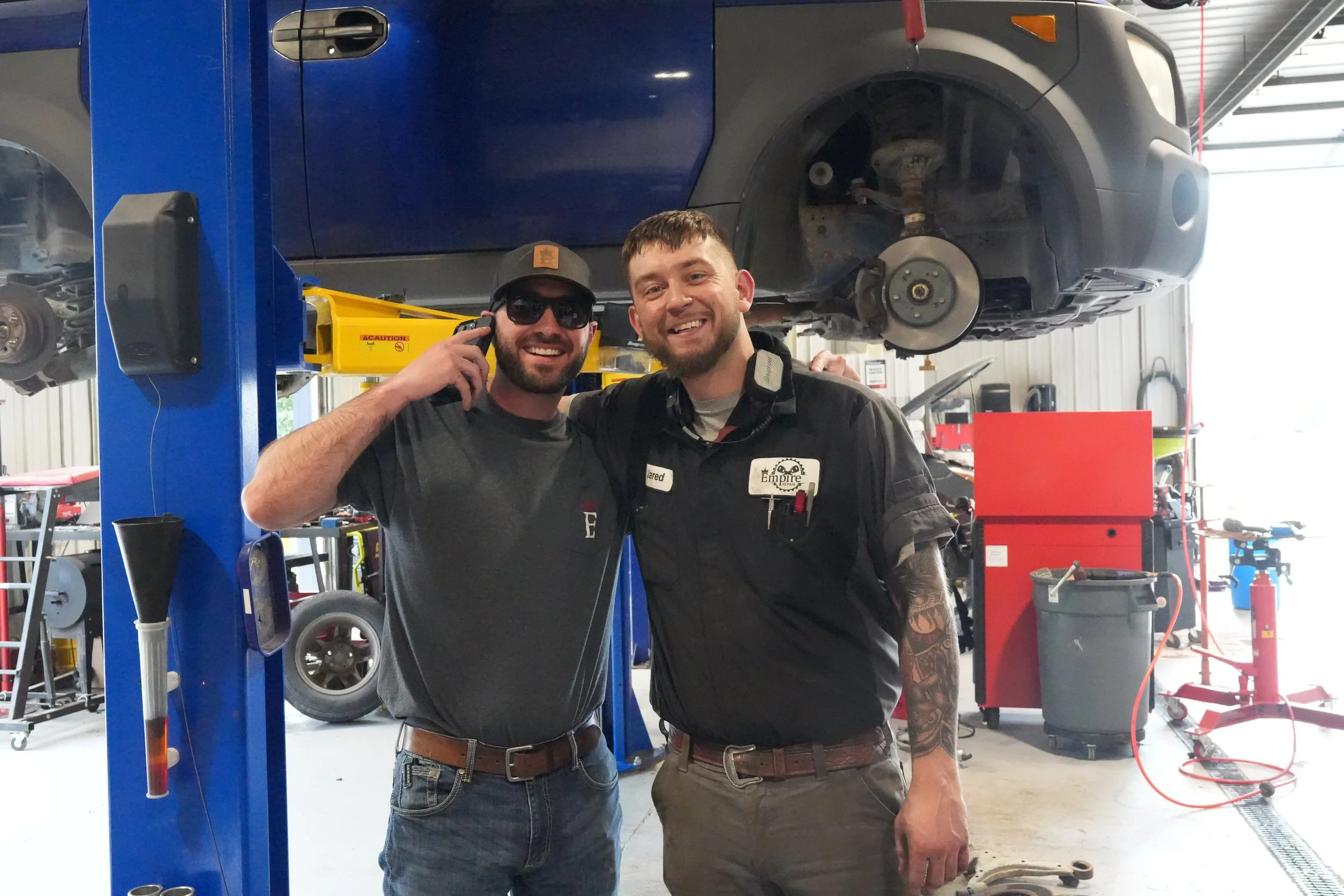 Two men smiling at the camera in an auto repair shop. One man is talking on the phone, wearing sunglasses and a dark cap; the other is wearing a repair uniform with a name tag. Behind them is a car lifted on a hydraulic lift with its wheel removed.