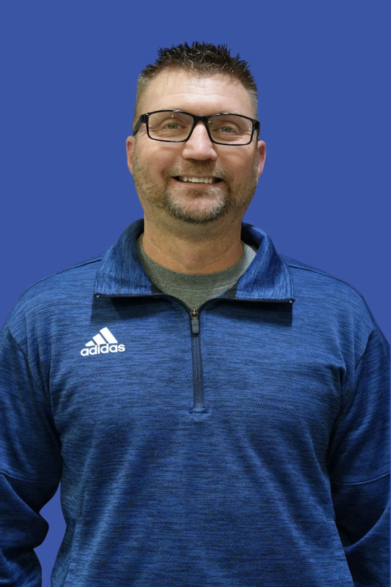 A man with short spiked hair, glasses, and a beard wearing a blue Adidas jacket, smiling, against a solid blue background.