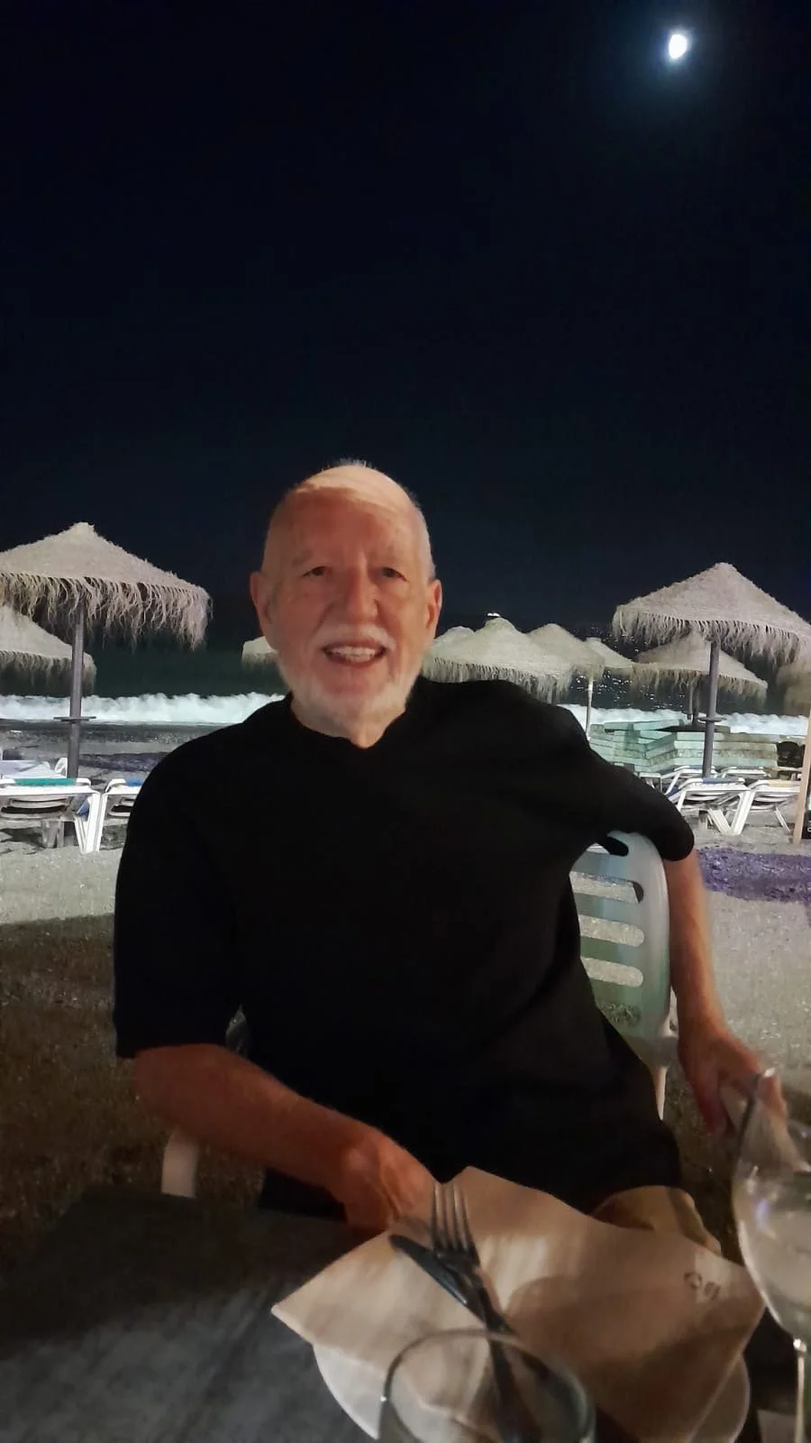 Hombre mayor sonriendo sentado en una mesa en la playa con sombrillas de paja y la luna en el cielo nocturno.