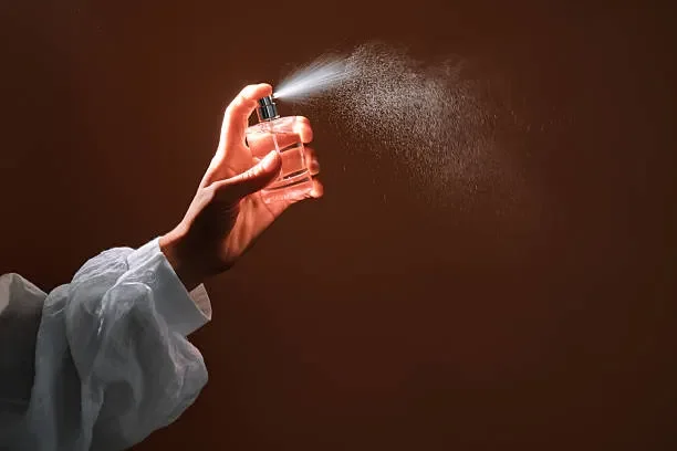A hand in a white sleeve holding a spray bottle dispensing mist against a brown background.