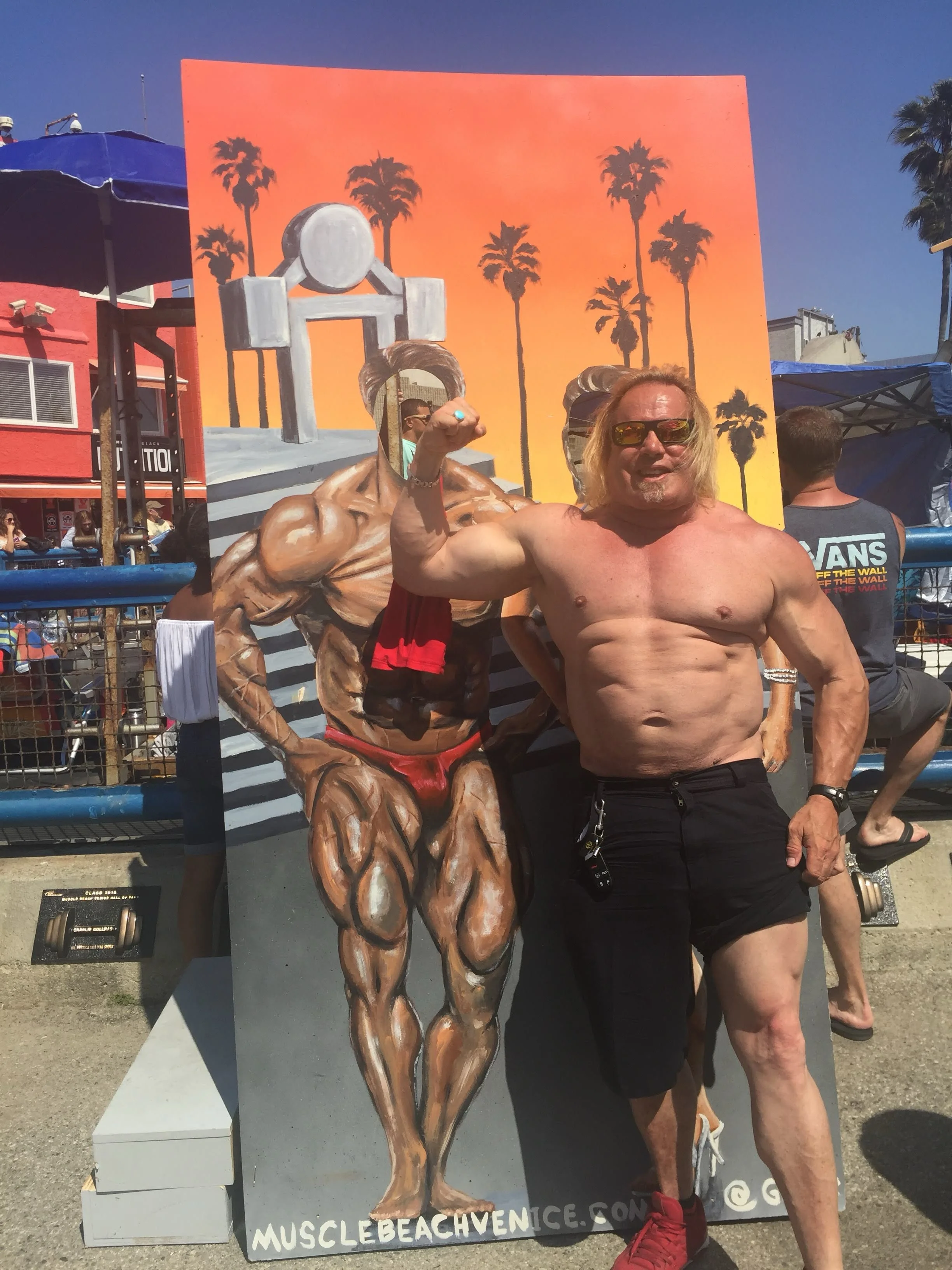 A shirtless man with blond hair and sunglasses standing next to a painted cutout of a muscular figure at the Muscle Beach Venice in Los Angeles. The background features an orange sunset sky with palm trees.