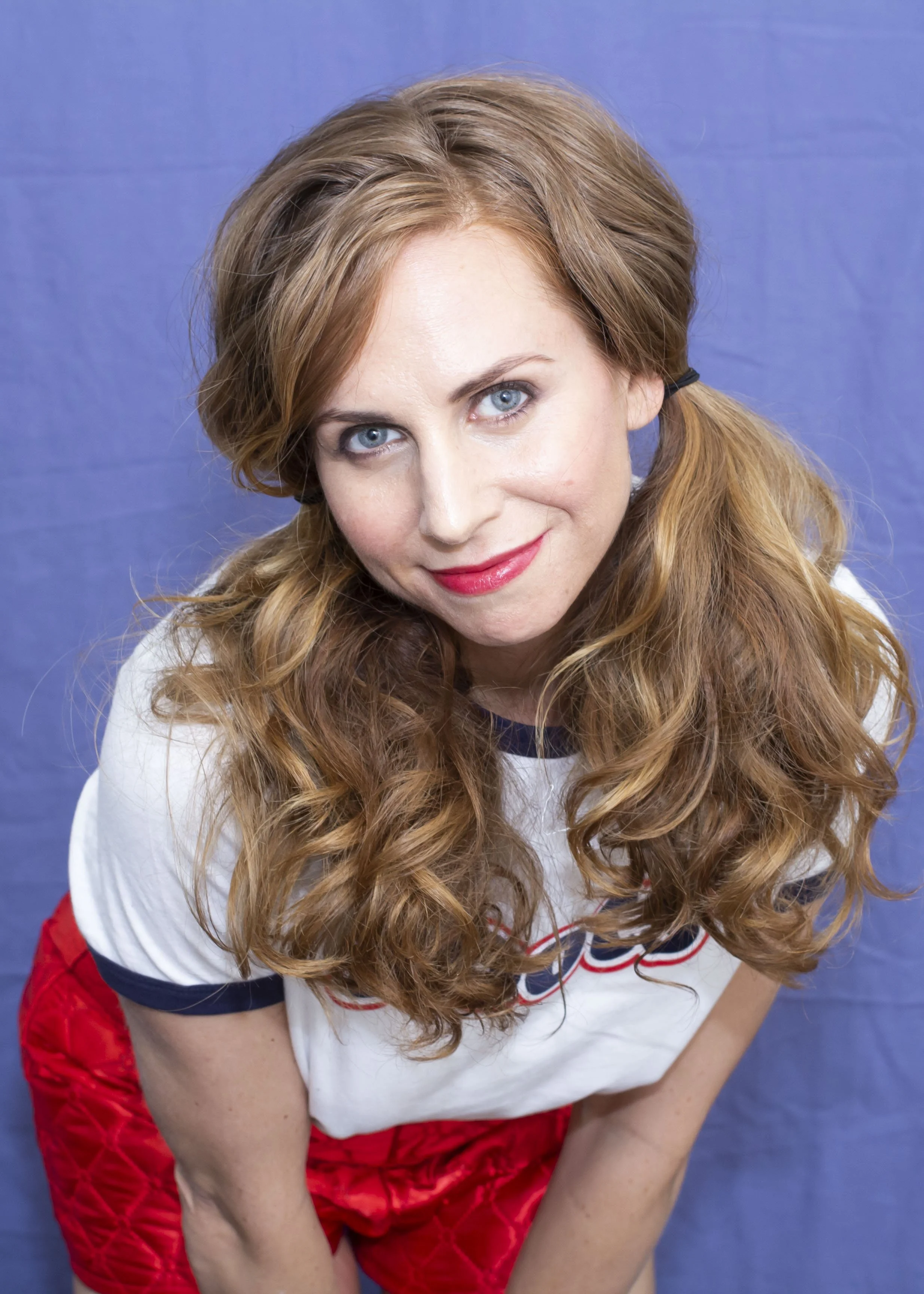 Jennifer Lieberman, red hair, blue eyes, and fair skin, wearing a white sports jersey with red and blue accents, and red shorts, standing against a blue background.