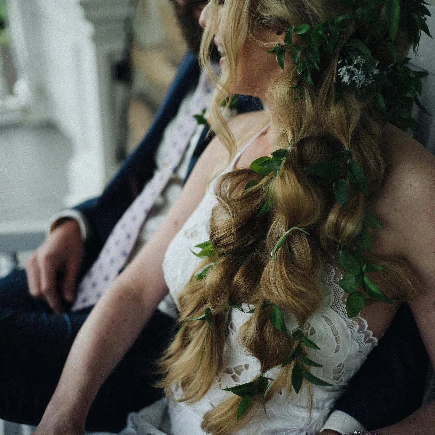 Close-up of a woman with long, curly blonde hair adorned with greenery, wearing a white lace top, sitting next to a man in a suit, at what appears to be a wedding or special ceremony.