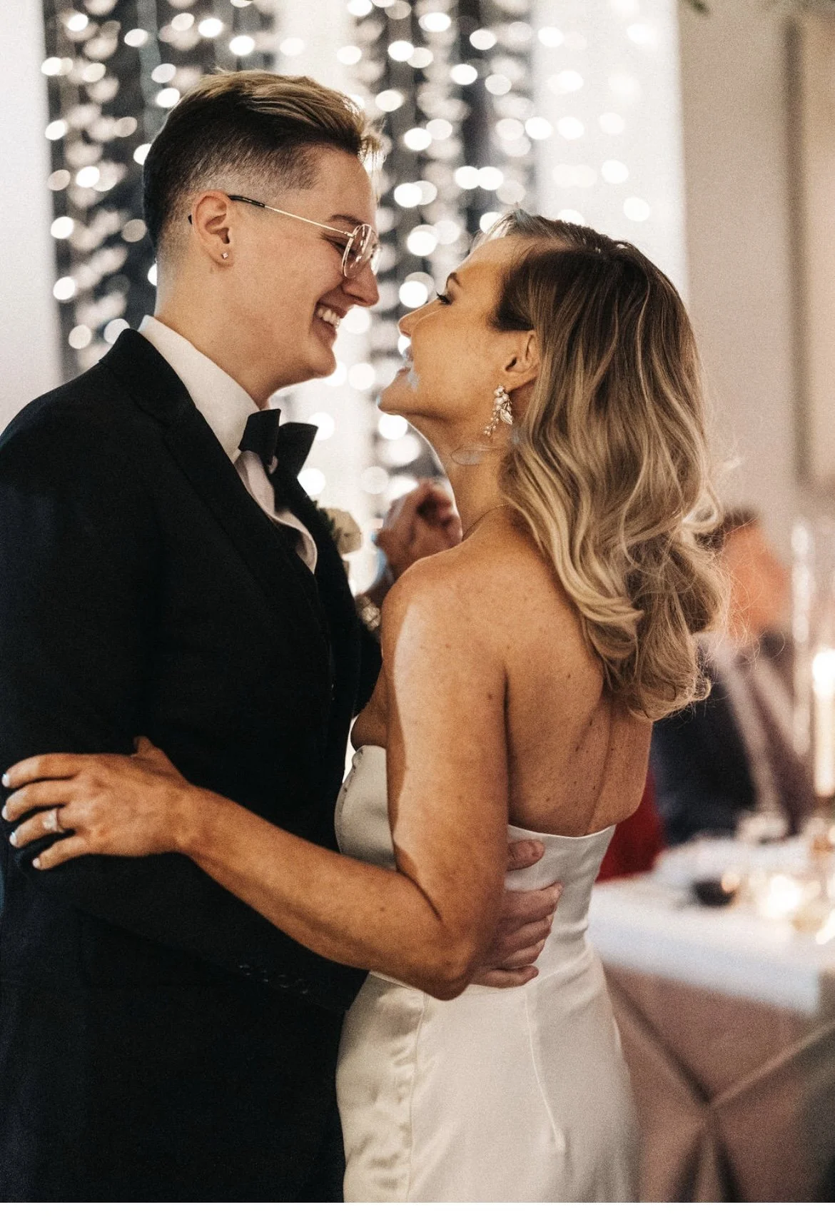 A couple dressed in wedding attire dancing and smiling at each other at their wedding reception, with a blurry background of guests and festive lights.
