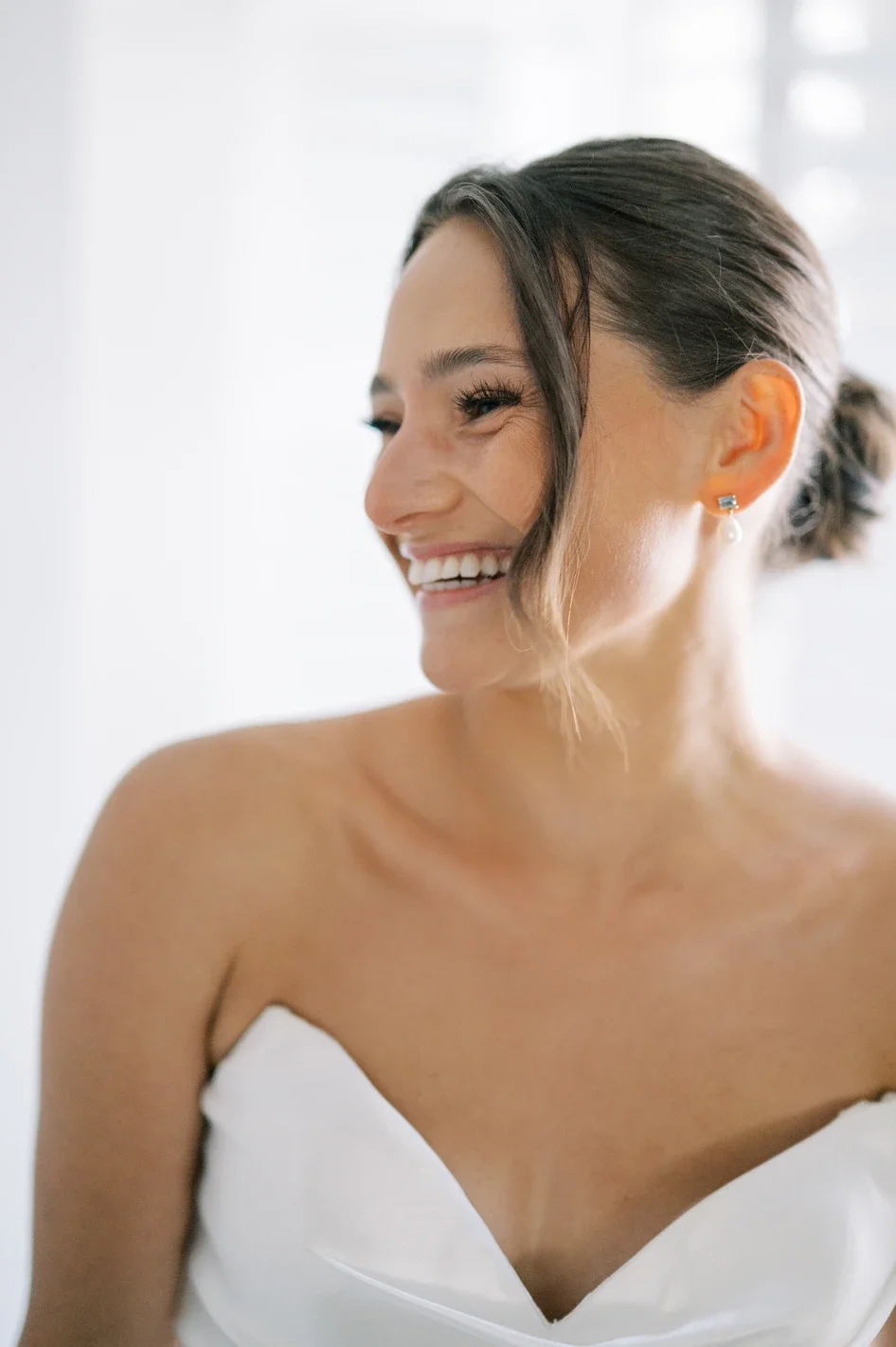 Smiling woman with dark hair styled in an updo, wearing pearl earrings and a white strapless dress, in a bright, airy room.