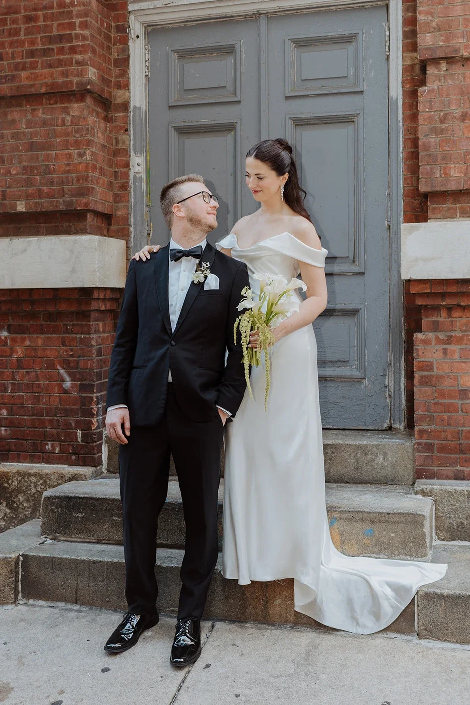 A bride and groom standing on the steps outside a building with a brick wall and gray door, looking at each other, the bride holding a bouquet of flowers.