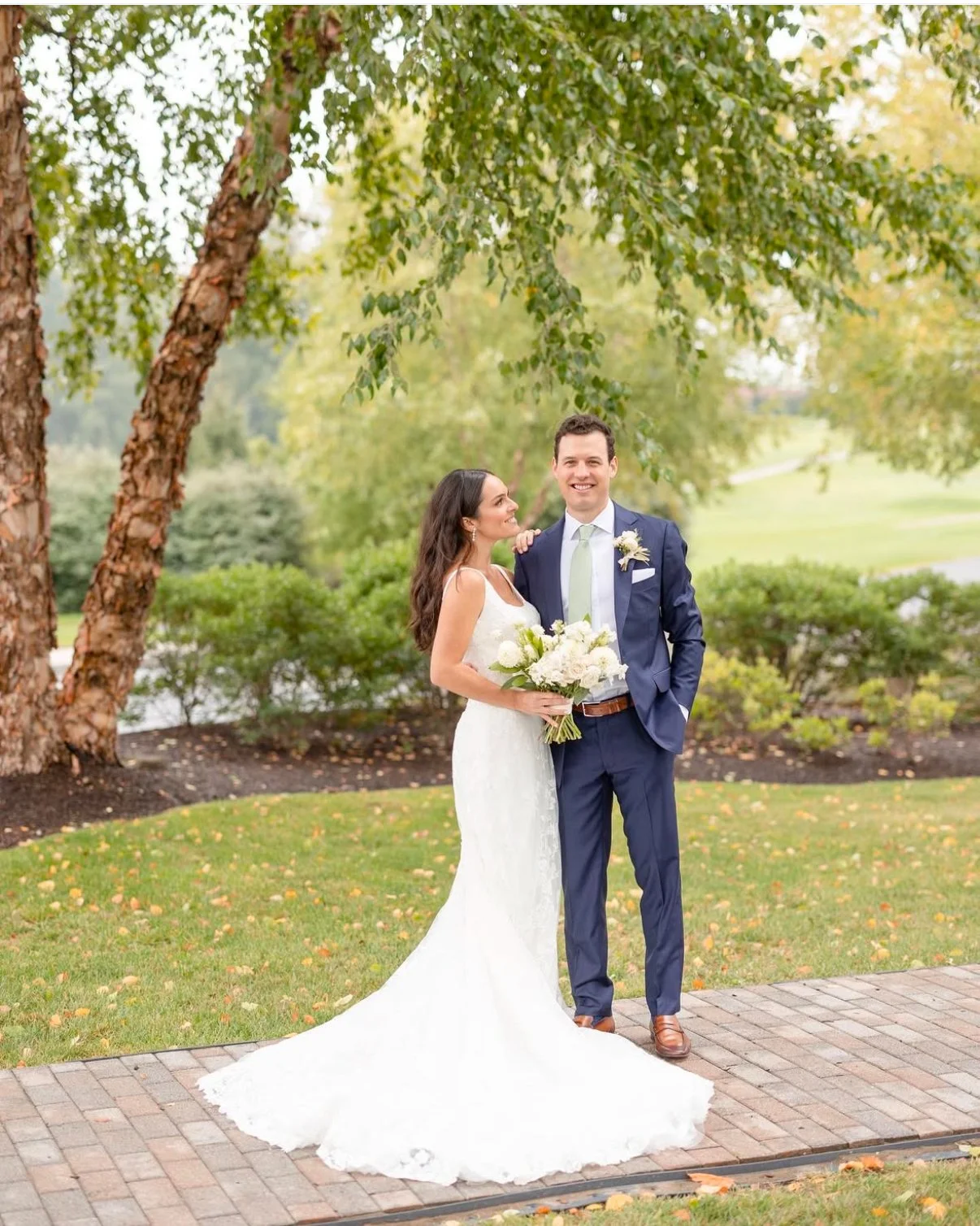 A bride and groom stand together outdoors on a brick pathway, smiling at each other. The bride is holding a bouquet of white flowers, wearing a white lace wedding dress with a long train, and has long dark hair. The groom is dressed in a navy blue su