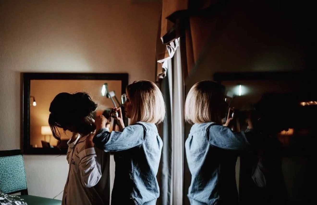 Two women in a hotel room, one applying makeup to the other, with their reflection seen in a mirror.