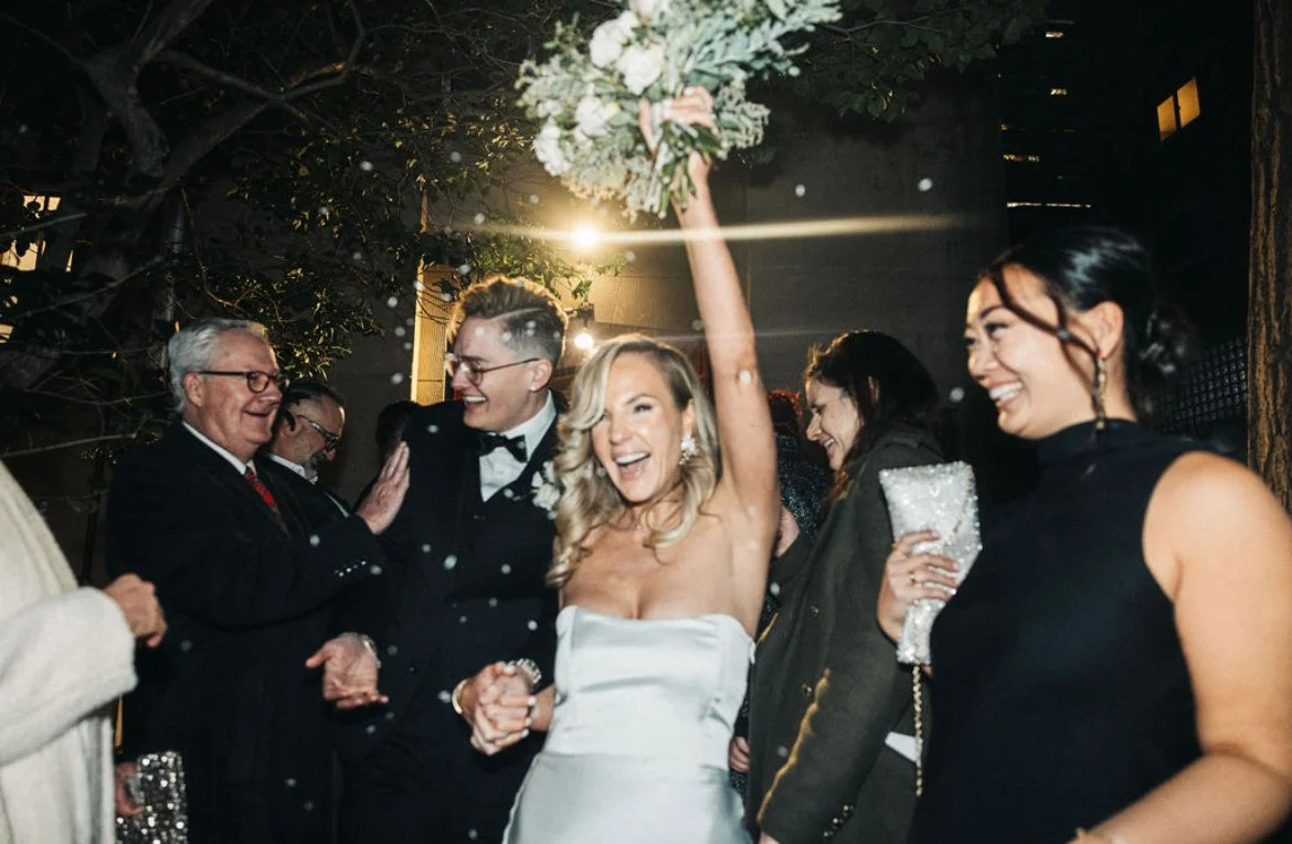 A bride in a white wedding dress holding a bouquet high while celebrating with cheerful guests at a wedding reception outdoor at night.