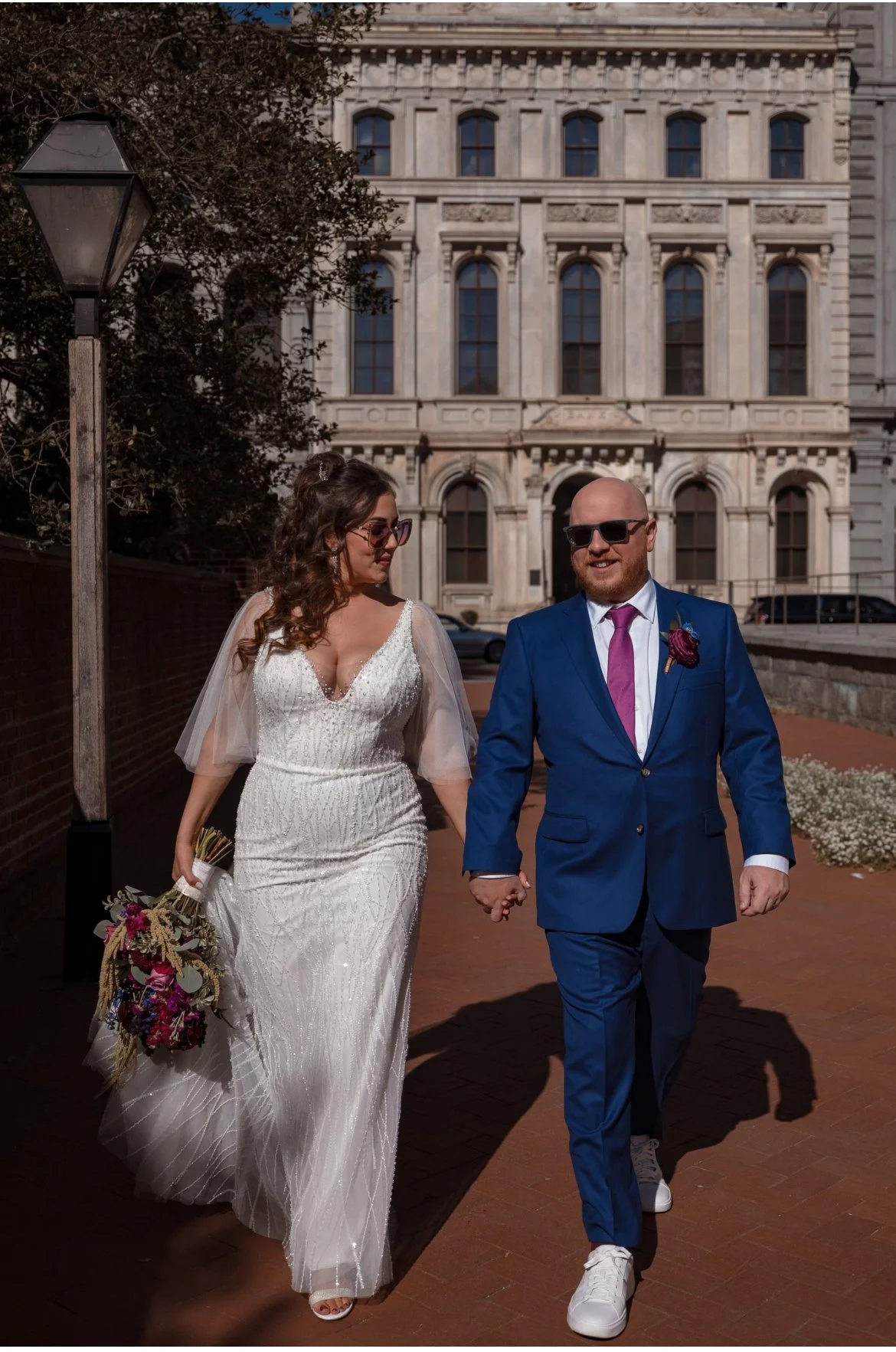 A bride and groom walking hand in hand outside a historic building, with the bride holding a bouquet of flowers and wearing a white wedding dress with a deep neckline, the groom in a blue suit with a pink tie, both wearing sunglasses.
