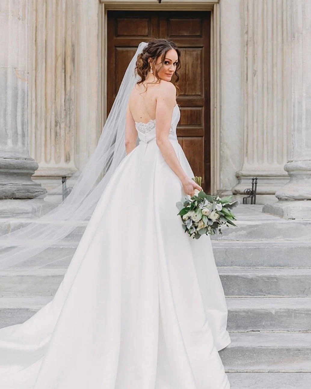 Bride in a white wedding gown holding a bouquet standing outside a building with a wooden door.