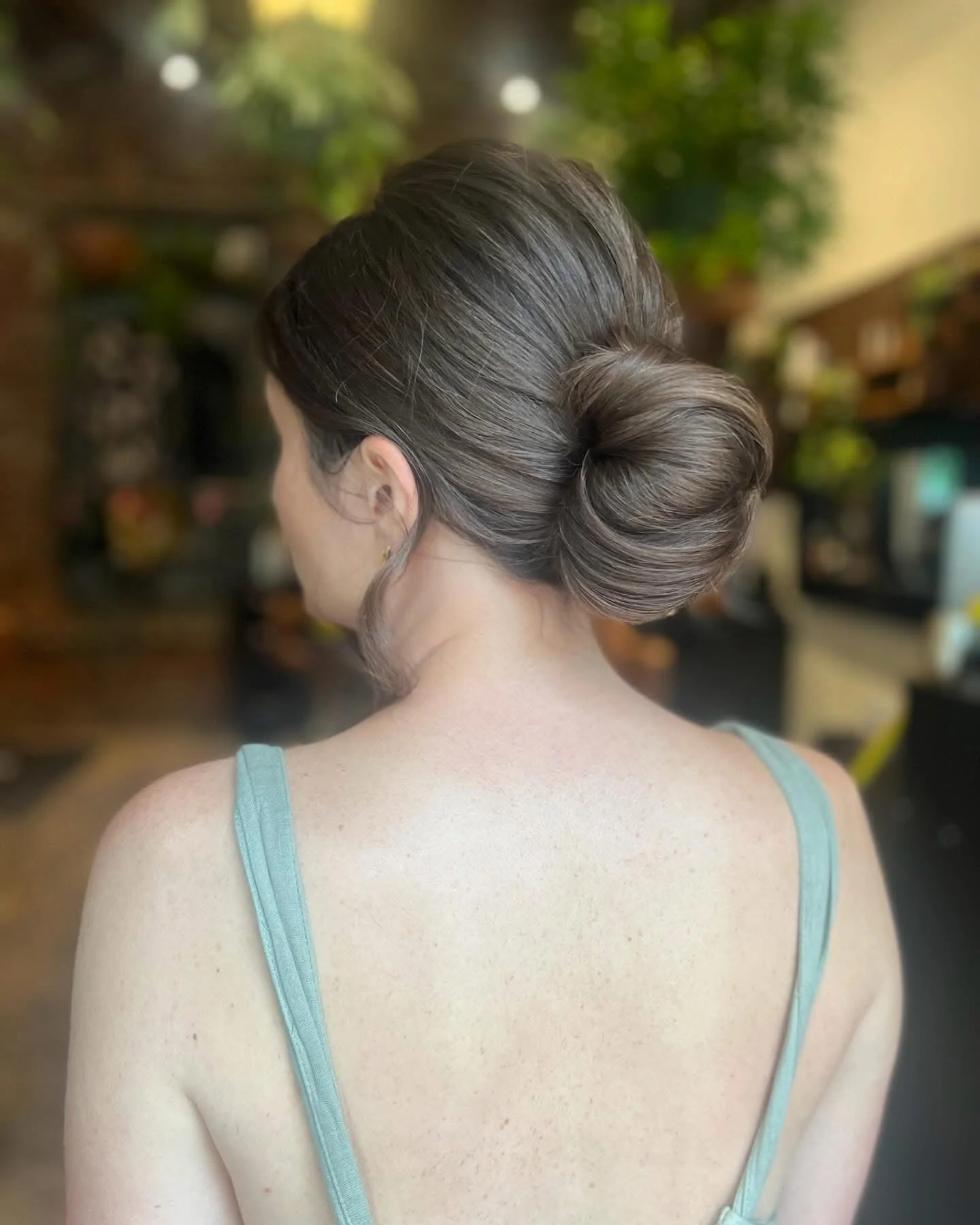 Woman with brown hair styled in an elegant updo, wearing a light blue tank top, standing indoors with blurred background of plants and furniture.