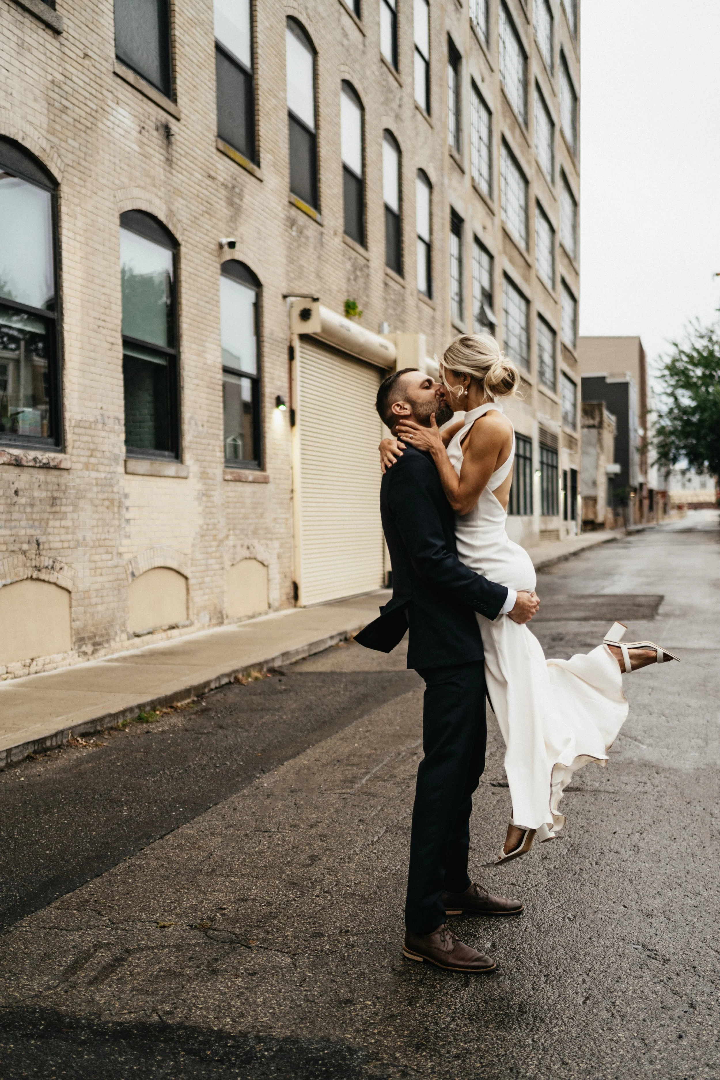 A man in a black suit lifting a woman in a white dress in front of a brick building, street scene, overcast sky.