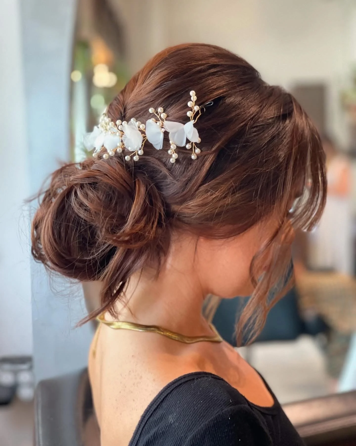 A woman with styled brown hair in loose curls, adorned with a decorative hairpiece with white flowers and pearls, wearing a black dress and a gold choker necklace.