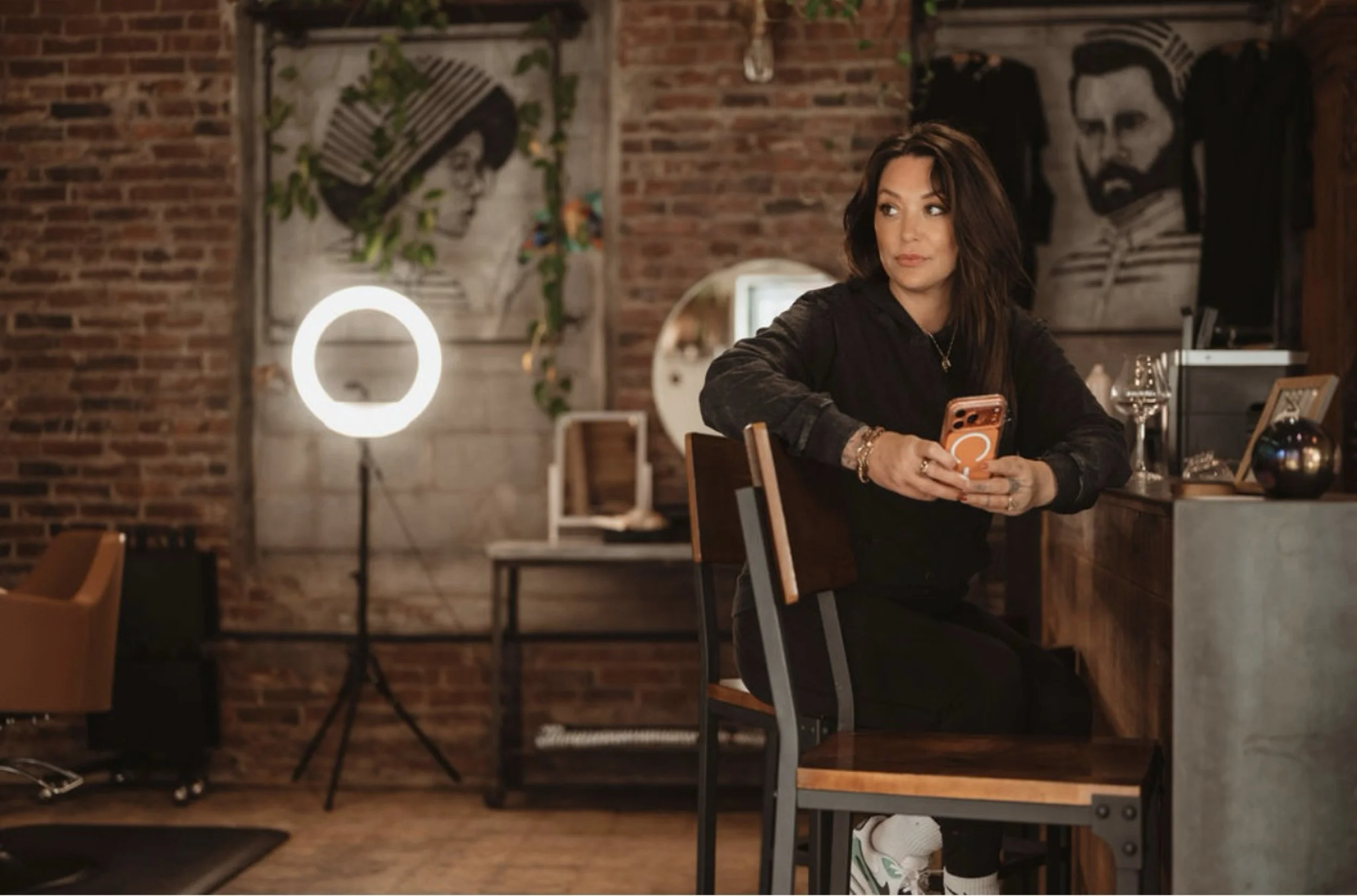 A woman with dark hair and a black top sitting at a table in a dimly lit restaurant with exposed brick walls and artwork, looking to her left while holding a smartphone.