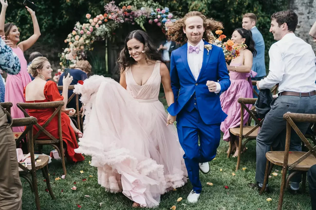 A newlywed couple, a woman in a pink wedding dress and a man in a blue suit, walking hand in hand down the aisle at an outdoor wedding ceremony with guests smiling and celebrating.
