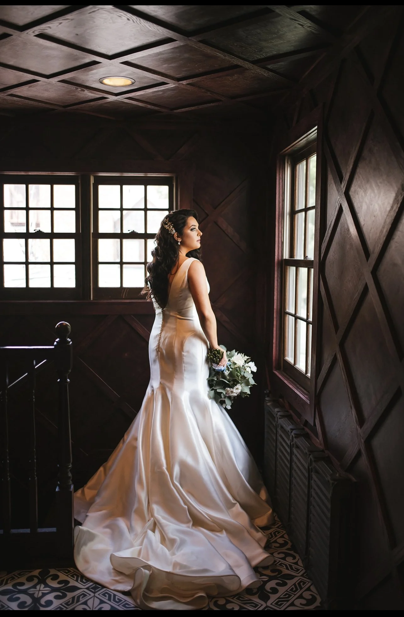 Bride standing by a window in a dimly lit room with dark wood paneling, wearing a satin wedding gown and holding a bouquet of white roses and greenery.