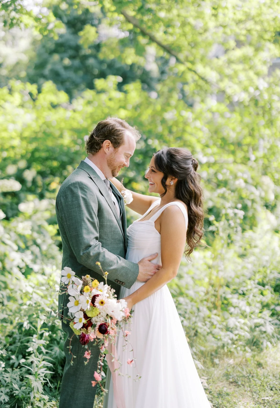 A bride and groom in wedding attire in a lush green outdoor setting, smiling at each other; the bride is holding a bouquet of flowers.