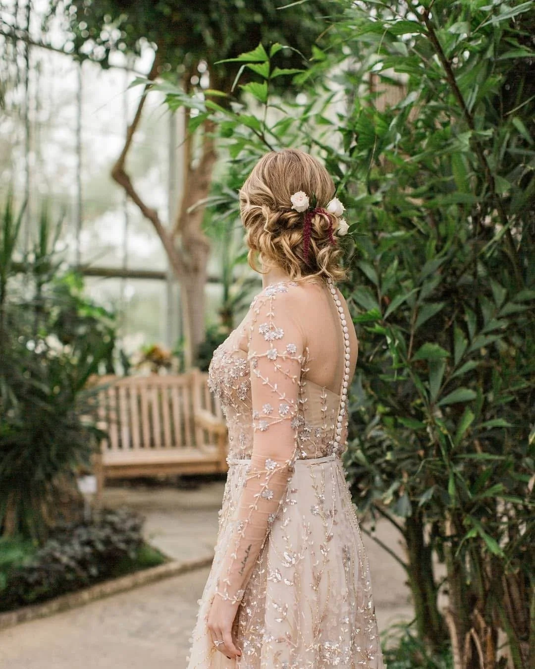 A woman in an elegant, embroidered gown with floral details, standing in a lush greenhouse with green plants and a wooden bench in the background.