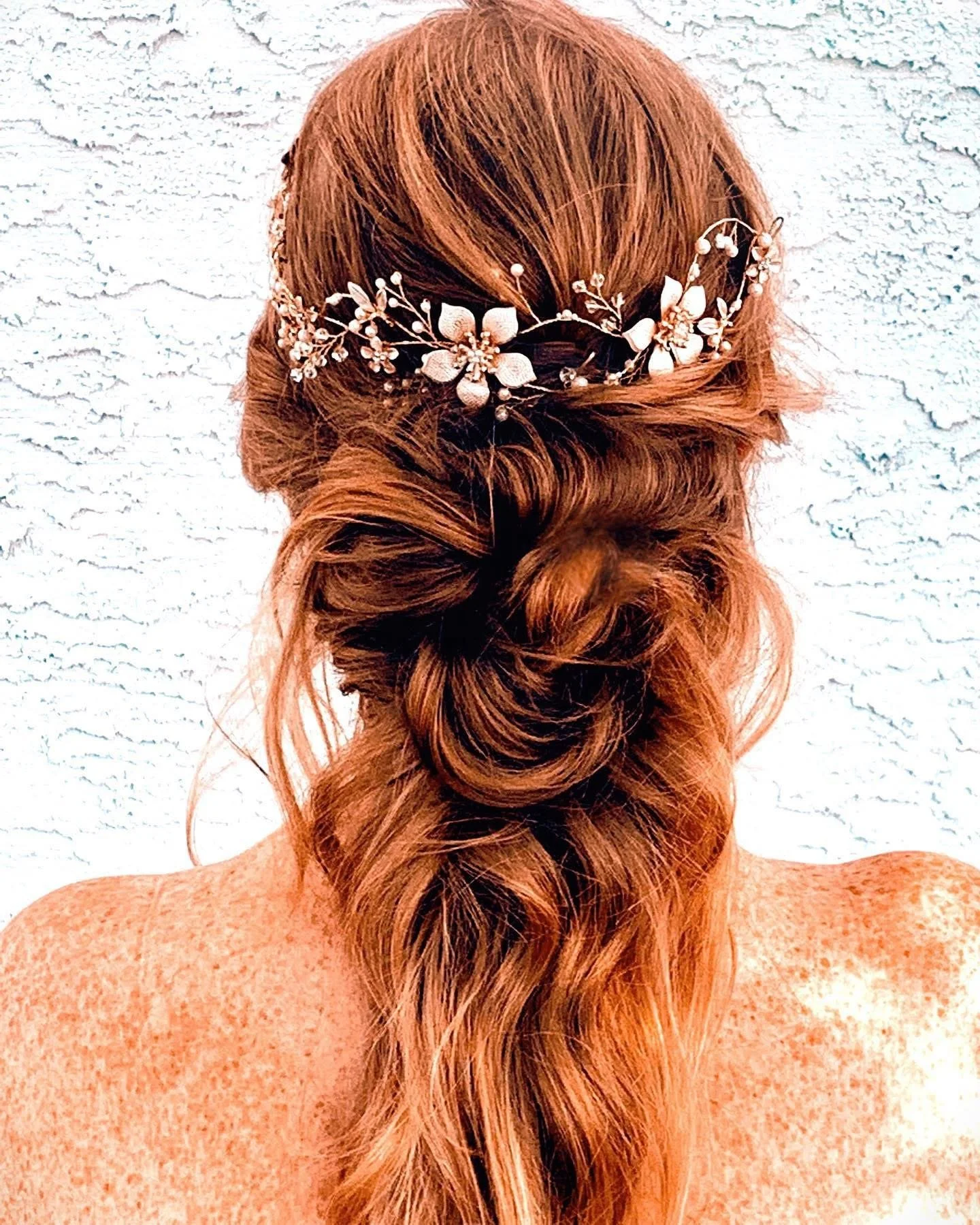 Back view of a woman with long red hair styled in loose waves and a braid, wearing a floral hair accessory, against a white textured wall.