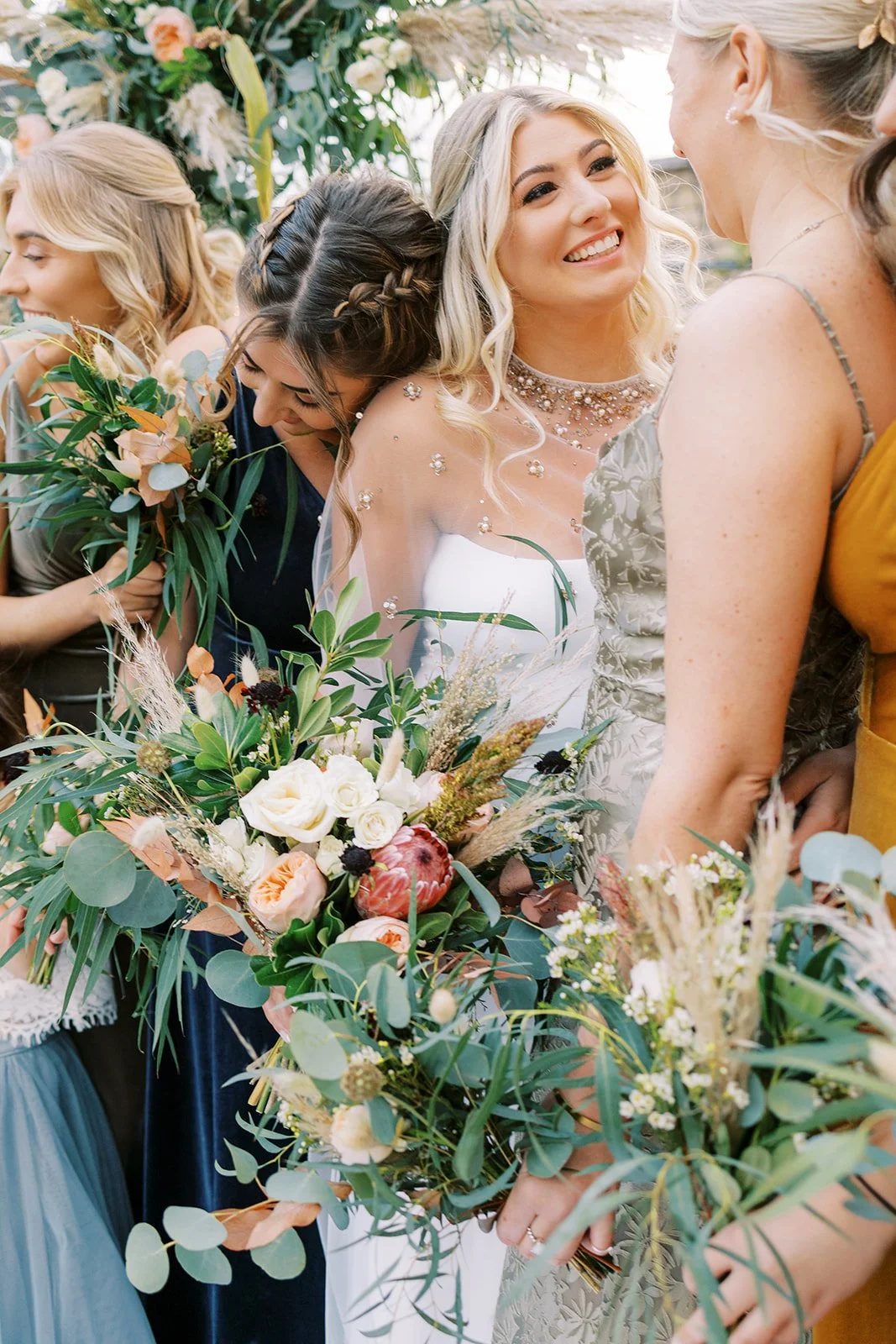 A bride with blonde hair smiling and wearing a strapless wedding dress with pearl embellishments, holding a large bouquet of flowers. She is surrounded by women, likely bridesmaids, dressed in colorful dresses, with one woman on the right engaging wi