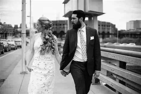 Philly Bride- A man and woman walking together outdoors in a city setting, holding hands and looking at each other, with buildings and a bridge in the background. Hair & Make-Up Mack Stylist 