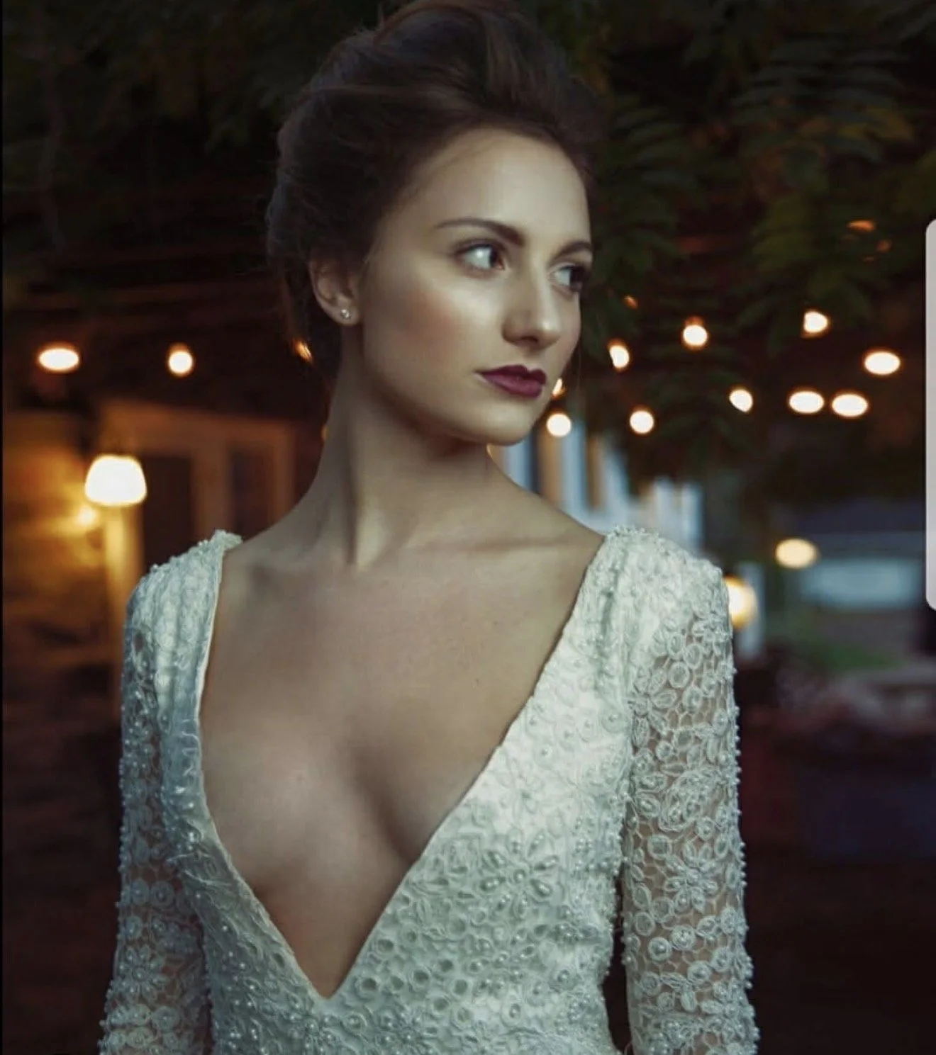 A young woman in a white lace wedding dress with a deep V neckline, looking to her left with an elegant hairstyle and dark lipstick. She is outdoors with blurred hanging lights and foliage in the background.