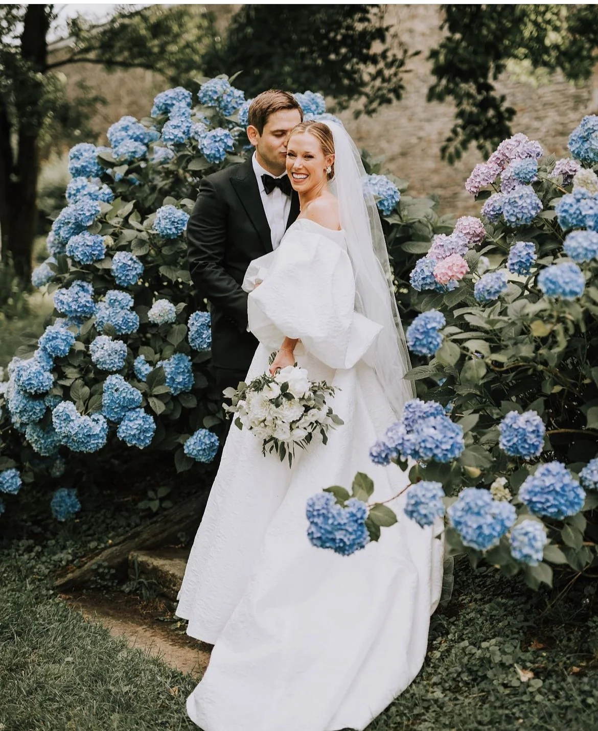 A bride and groom standing in front of a bush of blue and purple hydrangeas, smiling during their wedding photos. The groom is wearing a black tuxedo, and the bride is in a white wedding gown holding a bouquet of white flowers.