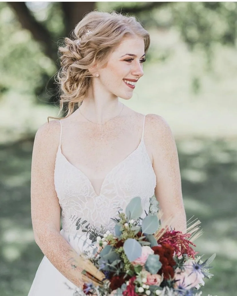 A smiling woman with wavy blonde hair in a wedding dress holding a bouquet of flowers outdoors.